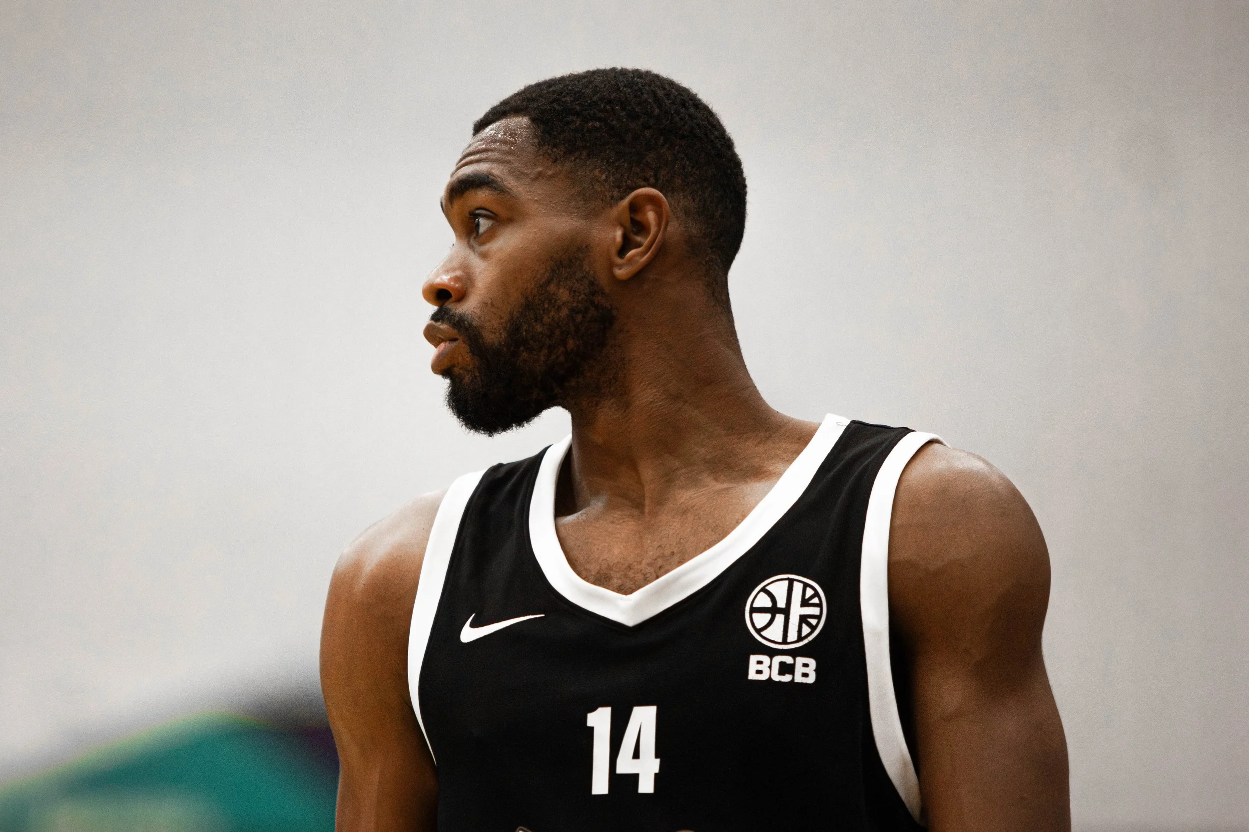 A male athlete in a black basketball jersey with the number 14, beard, and short hair looking to the side.