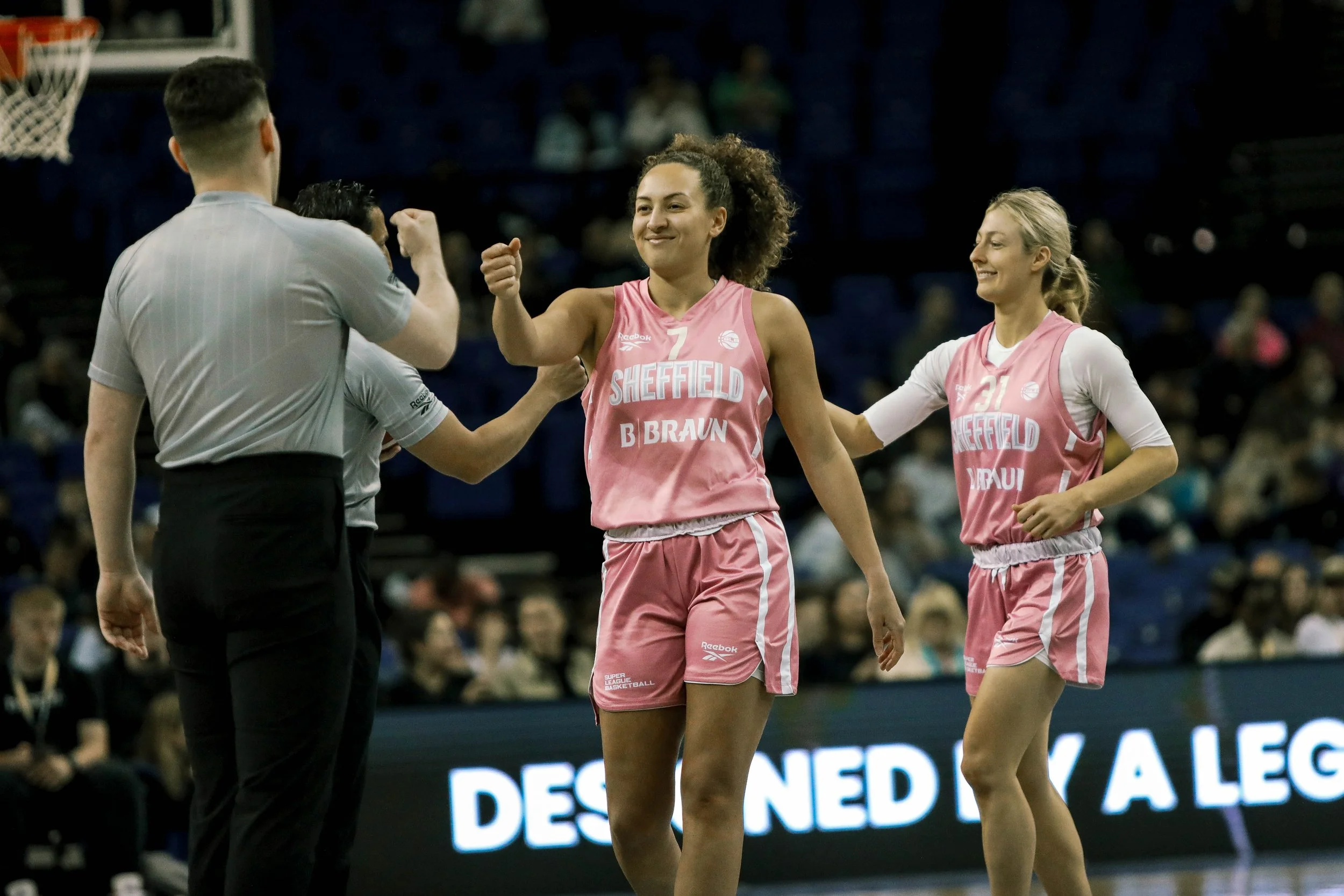 Two female basketball players in pink jerseys celebrate and greet the referee during a game, with one player giving a fist bump and the other smiling.