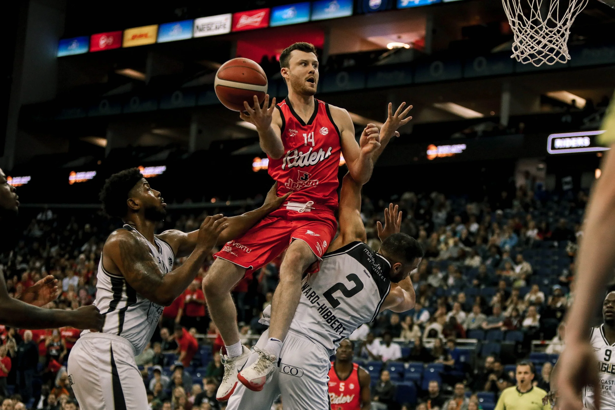 A basketball player in a red jersey with number 14 is jumping with the ball, attempting a shot while being defended by two players in white jerseys in an indoor basketball arena filled with spectators.