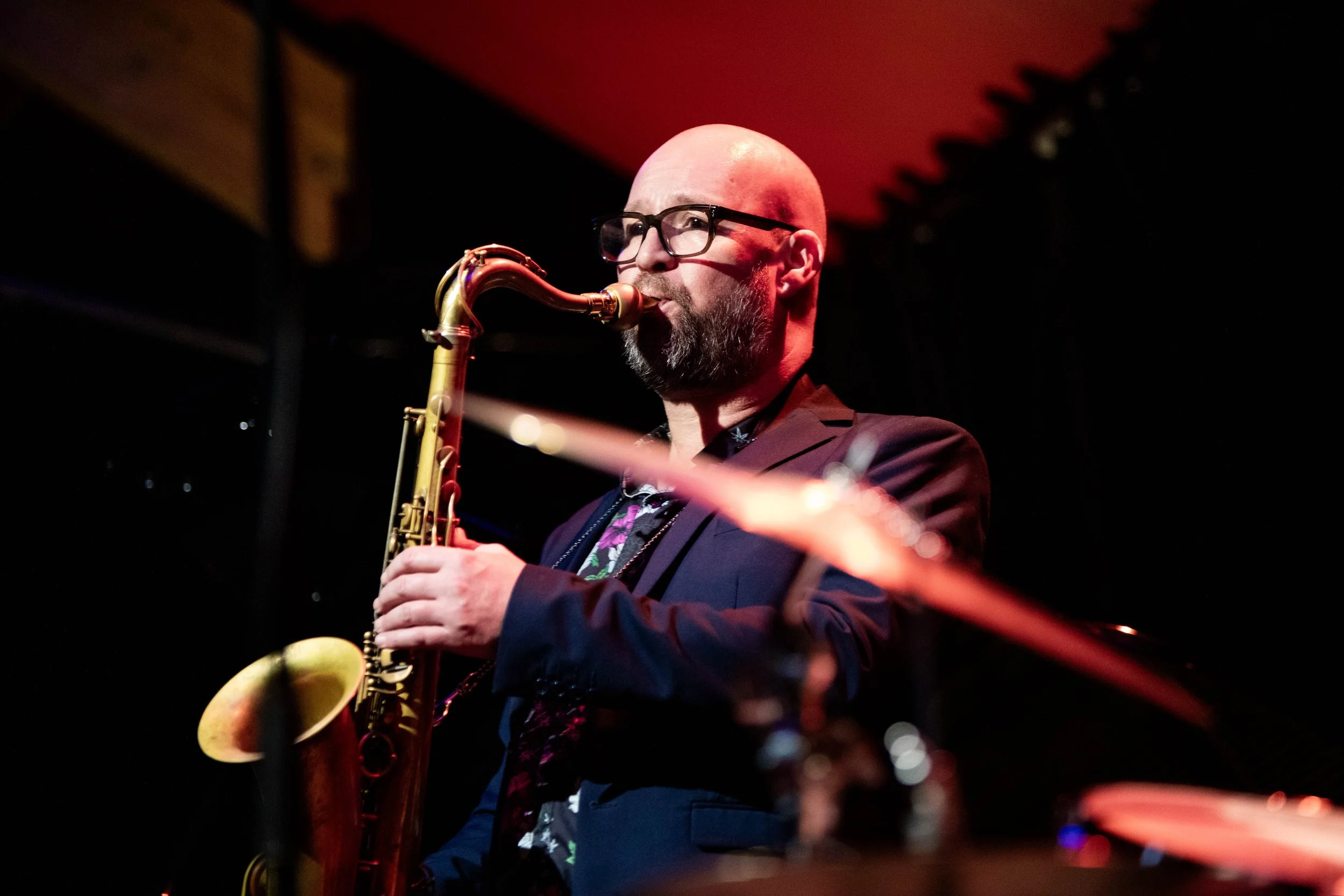 A man with glasses and a beard playing a saxophone on stage with red lighting.