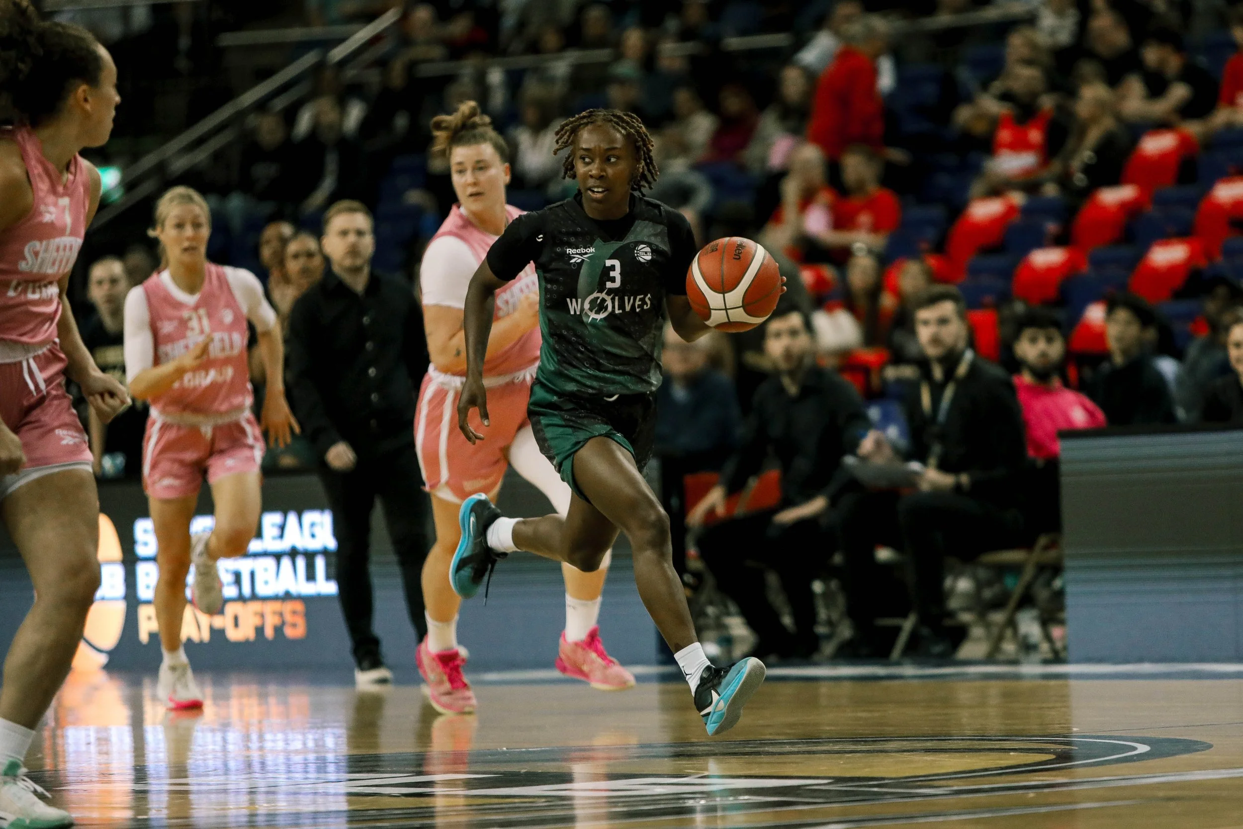 A female basketball player from the Wolves team dribbling the ball on the court during a game against a team wearing pink uniforms, with other players and spectators visible in the background.