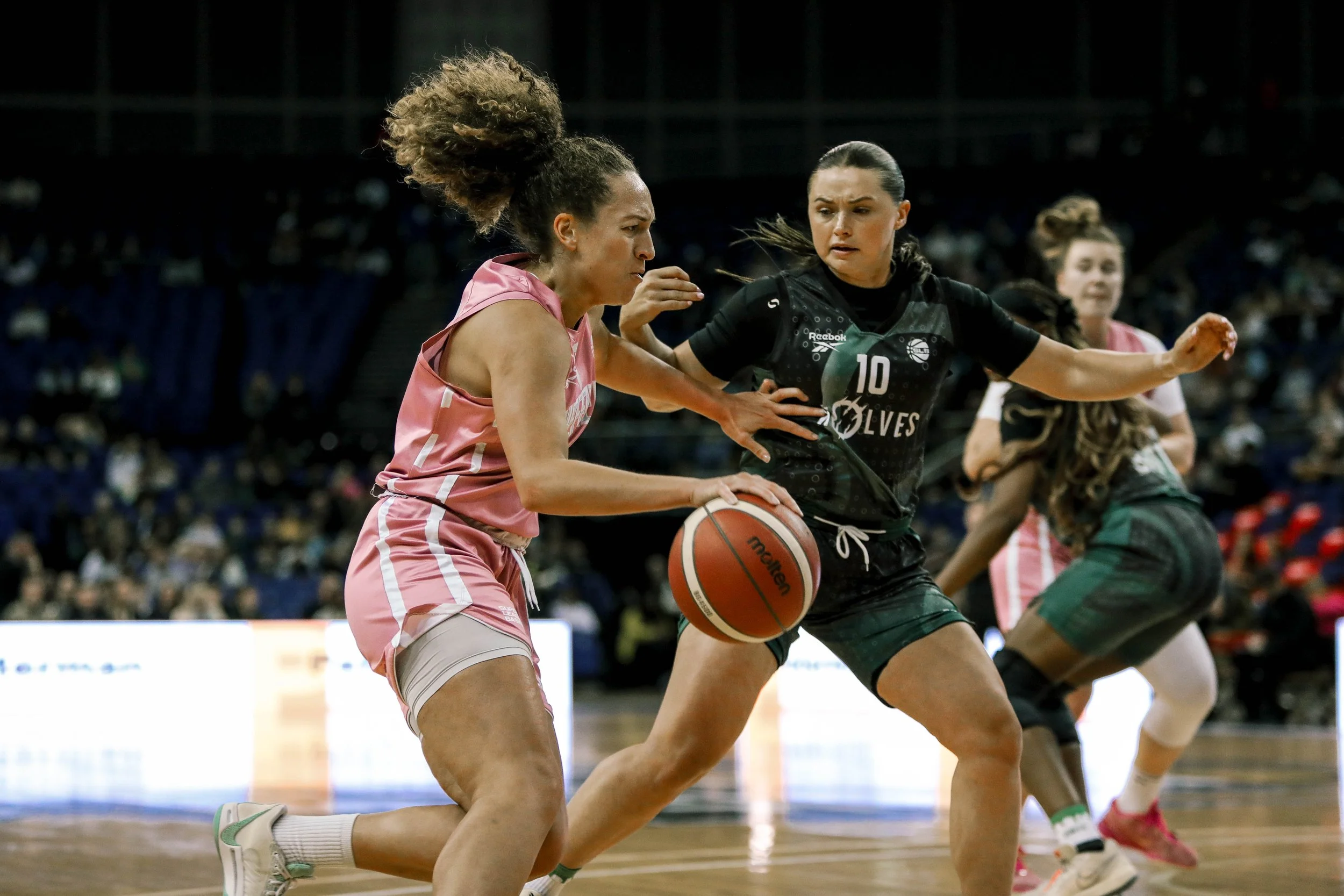 Women playing basketball in a game, with one player dribbling and being guarded by another.