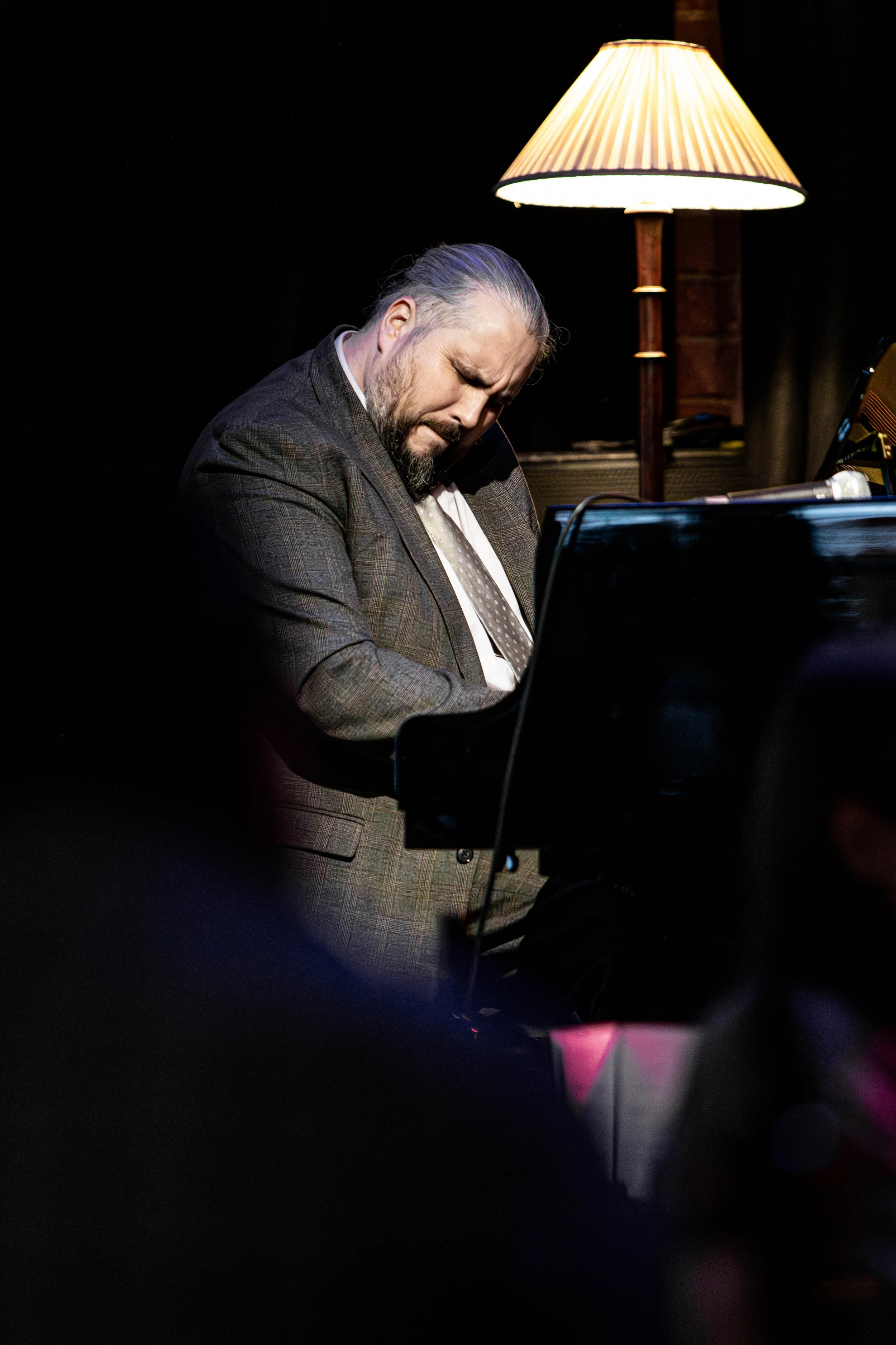 A man in a plaid suit playing the piano with a focused expression, positioned near a table lamp in a dimly lit space.