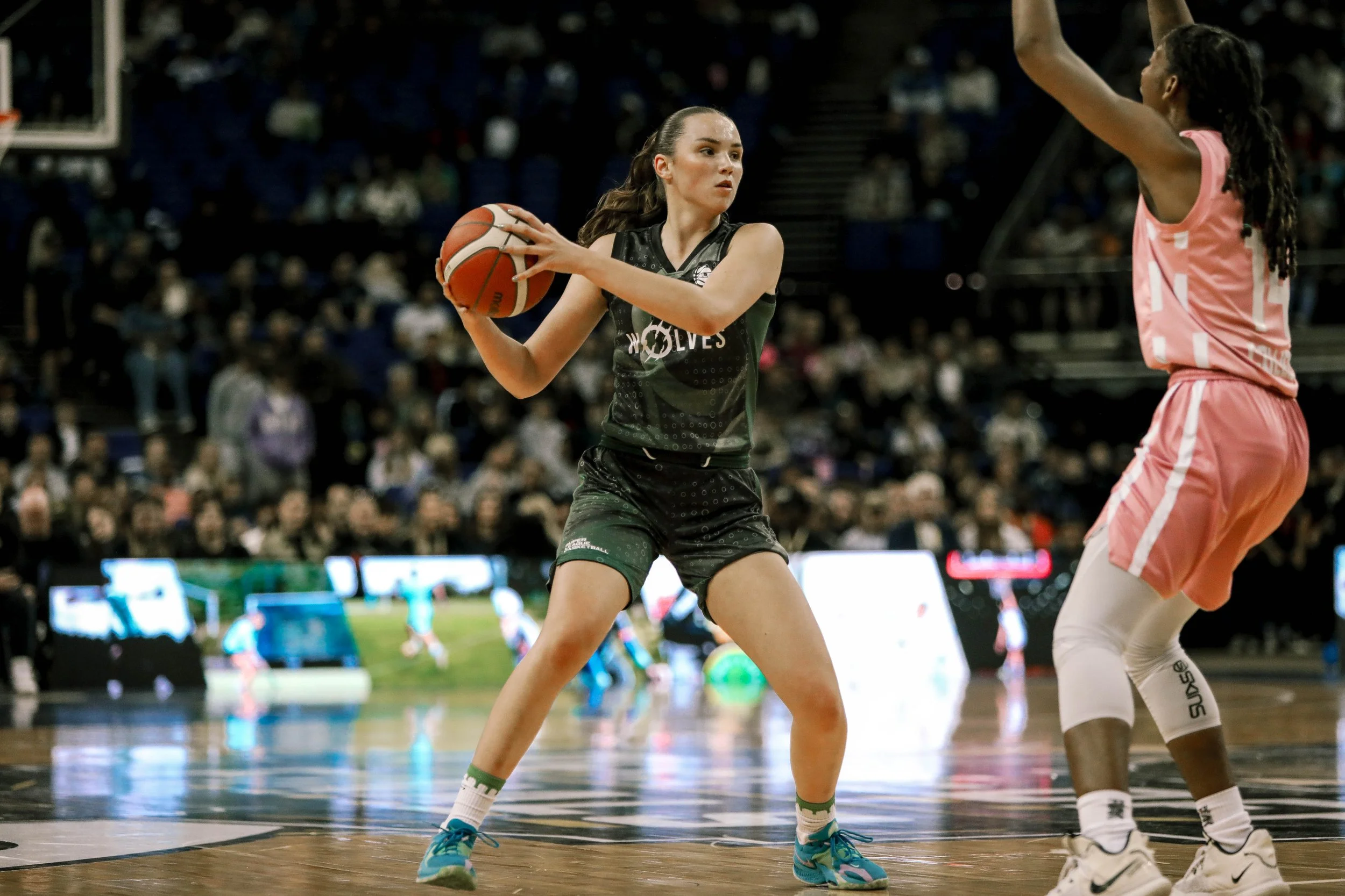 Two female basketball players are competing on the court during a game. The player in the black uniform holds the basketball and prepares to pass or shoot, while the player in the pink uniform attempts to block or defend. Spectators are visible in th