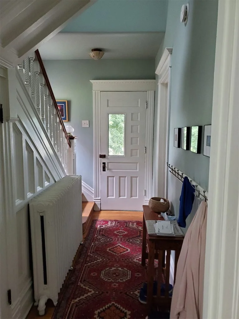 A narrow entryway with a white front door, a small window, and a staircase on the left. There's a red patterned runner rug on the wooden floor, a radiator, a small wooden table with newspapers and a basket, and a wall-mounted coat hook with jackets. 