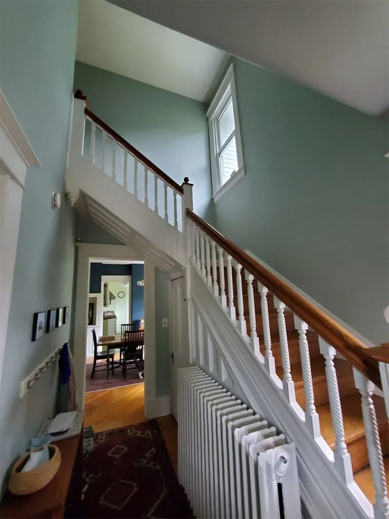 Interior view of a house showing a staircase with a wooden handrail, white spindles, and a radiator. Beyond the staircase is a dining area with a wooden table and chairs, partly visible through an open doorway. The walls are painted light blue, and a