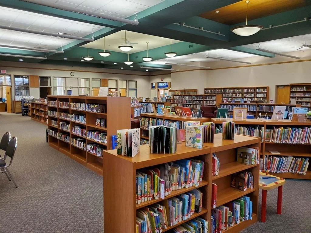 A section of a public library with wooden bookshelves filled with children's and adult books, chairs, and a large window in the background.