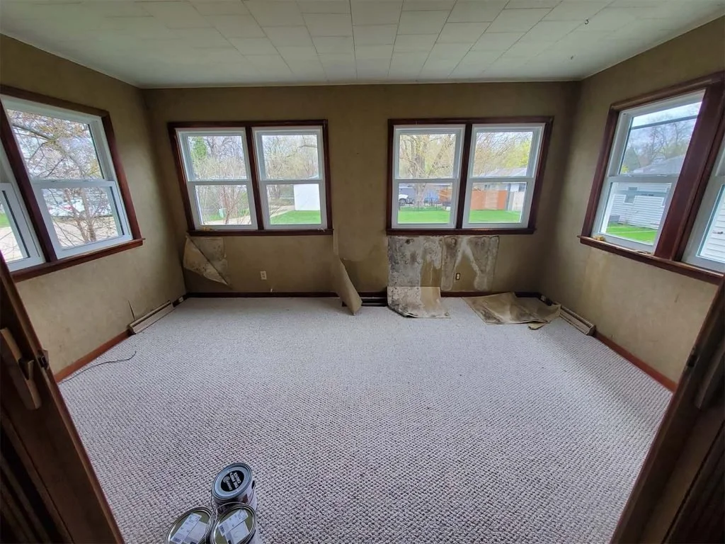 Room with multiple windows, walls with peeling wallpaper or paint, and carpeted flooring. Some torn paper or wallpaper on the lower part of the walls and a remote control or electronic device on the floor.