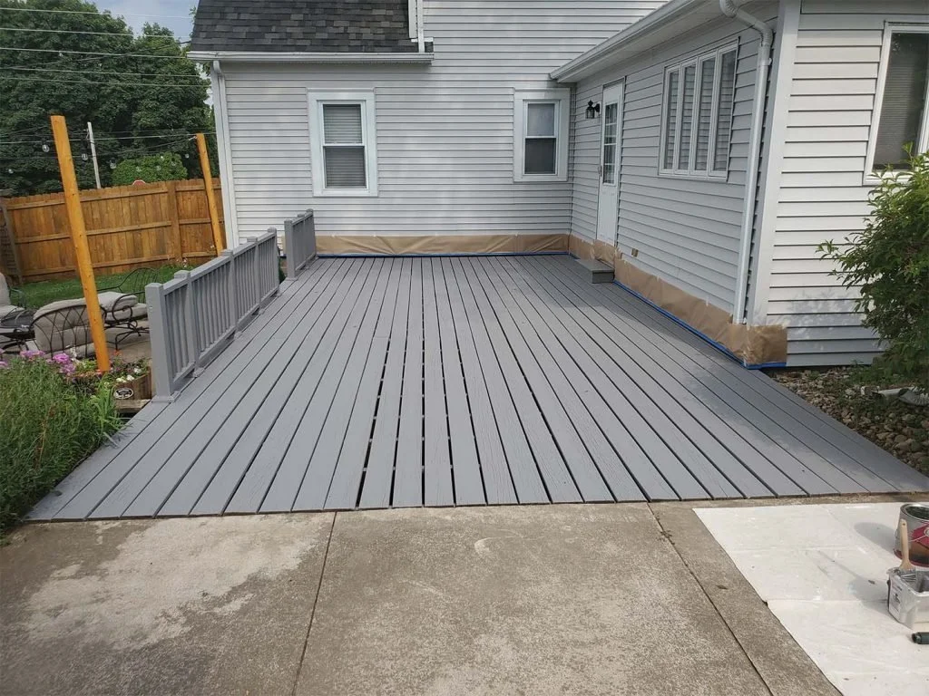 A new gray wooden deck attached to a house with white siding, partially covered with painter's paper, and an adjacent concrete patio with painting supplies.