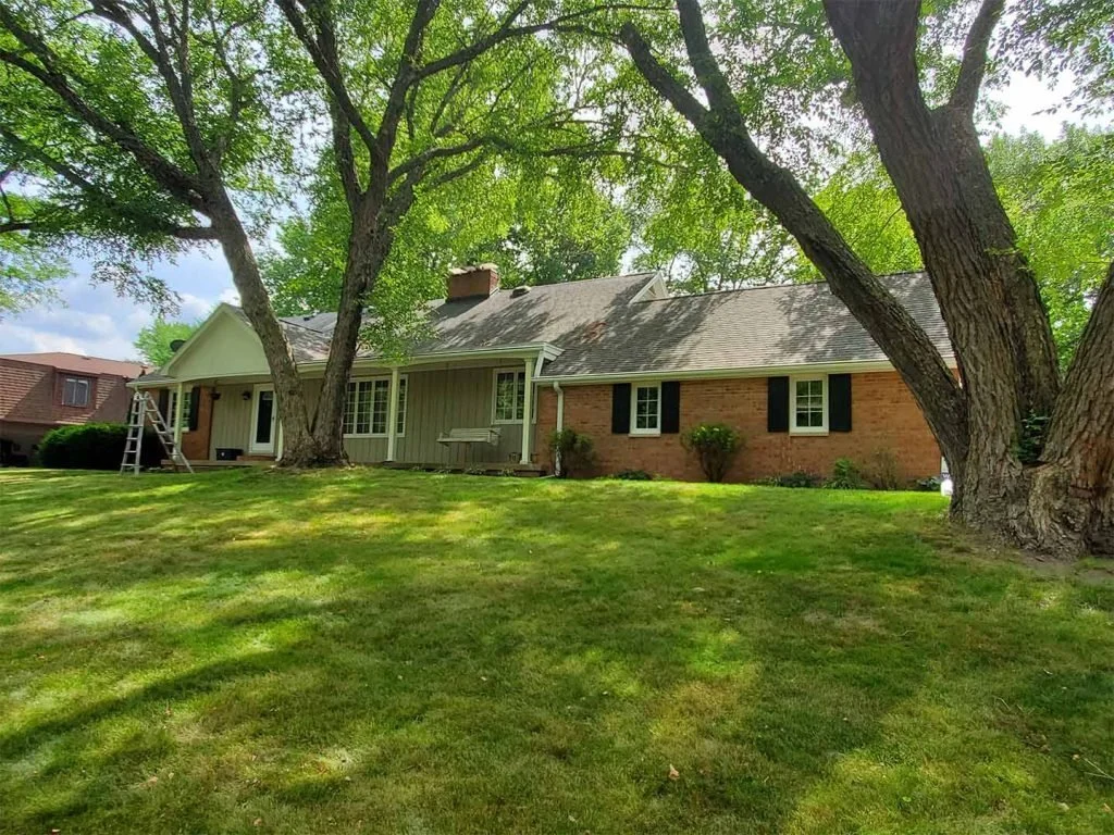 A house with brick and siding exterior, surrounded by large trees with lush green grass, some bushes, and a ladder leaning against the house.
