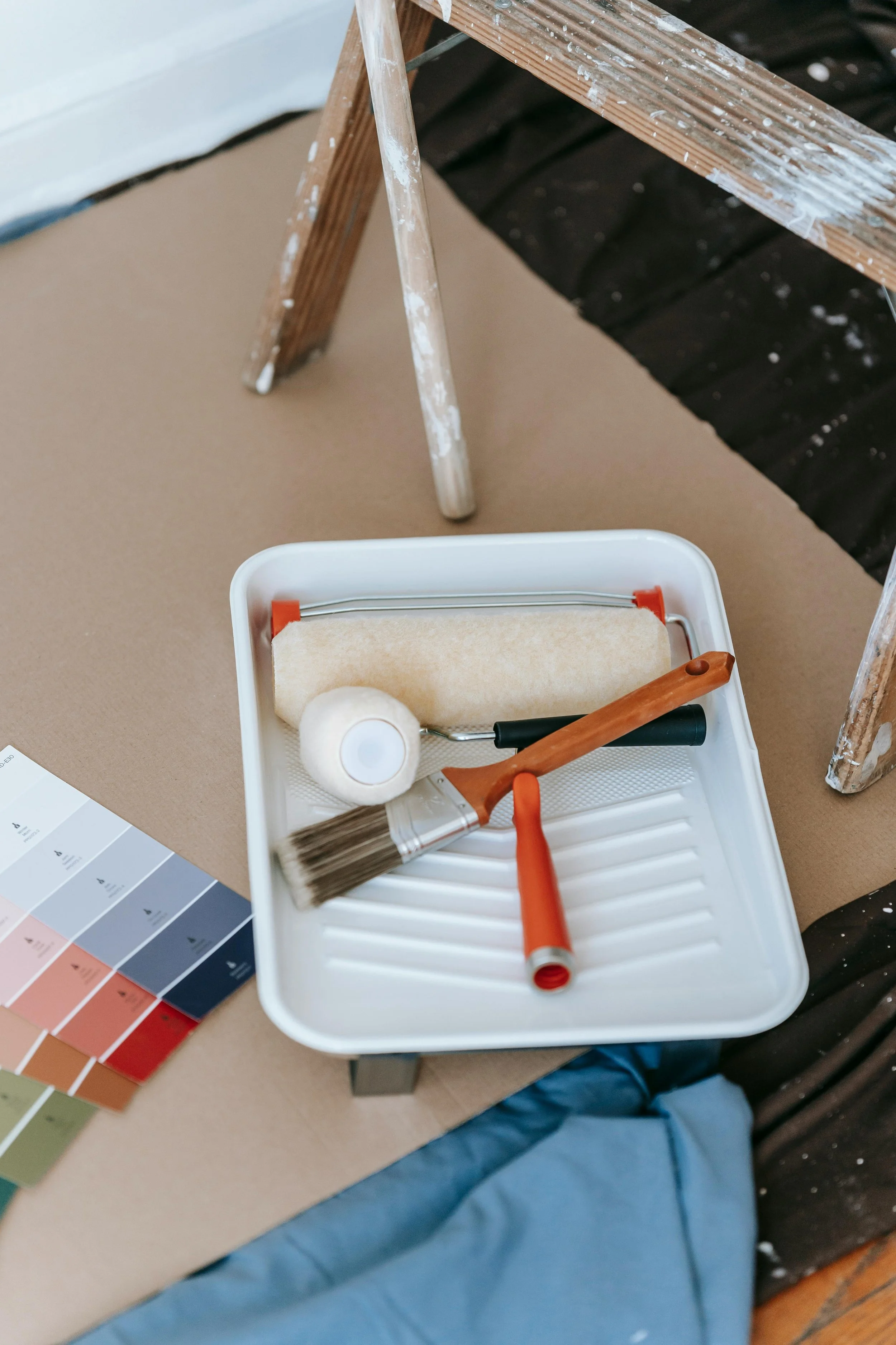 Painting tools including a paint roller, small paintbrush, roller handle, and a paint tray with a beige paint roller cover, on a table next to a color swatch.