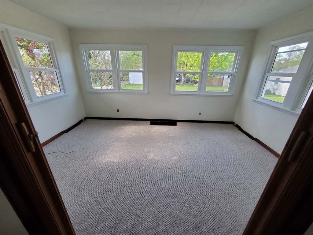 Empty room with white walls, large windows, and beige carpet, showing outdoor greenery and neighboring houses.