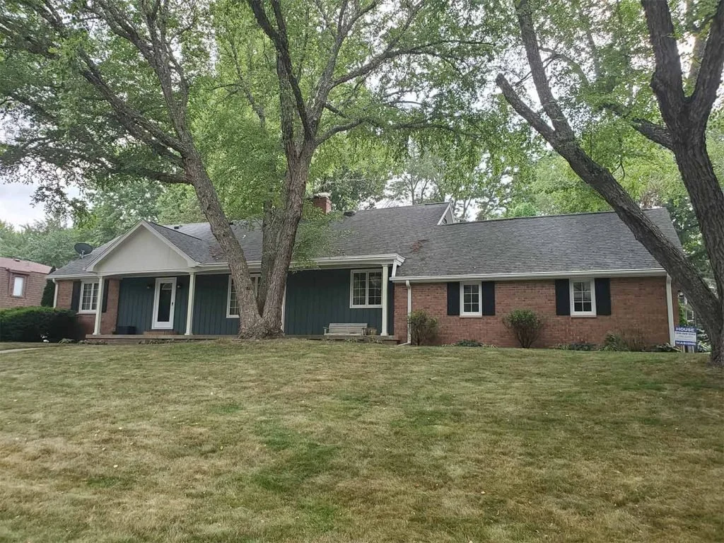 Suburban house with brick and blue siding, large trees in front, and a lawn with a sidewalk.