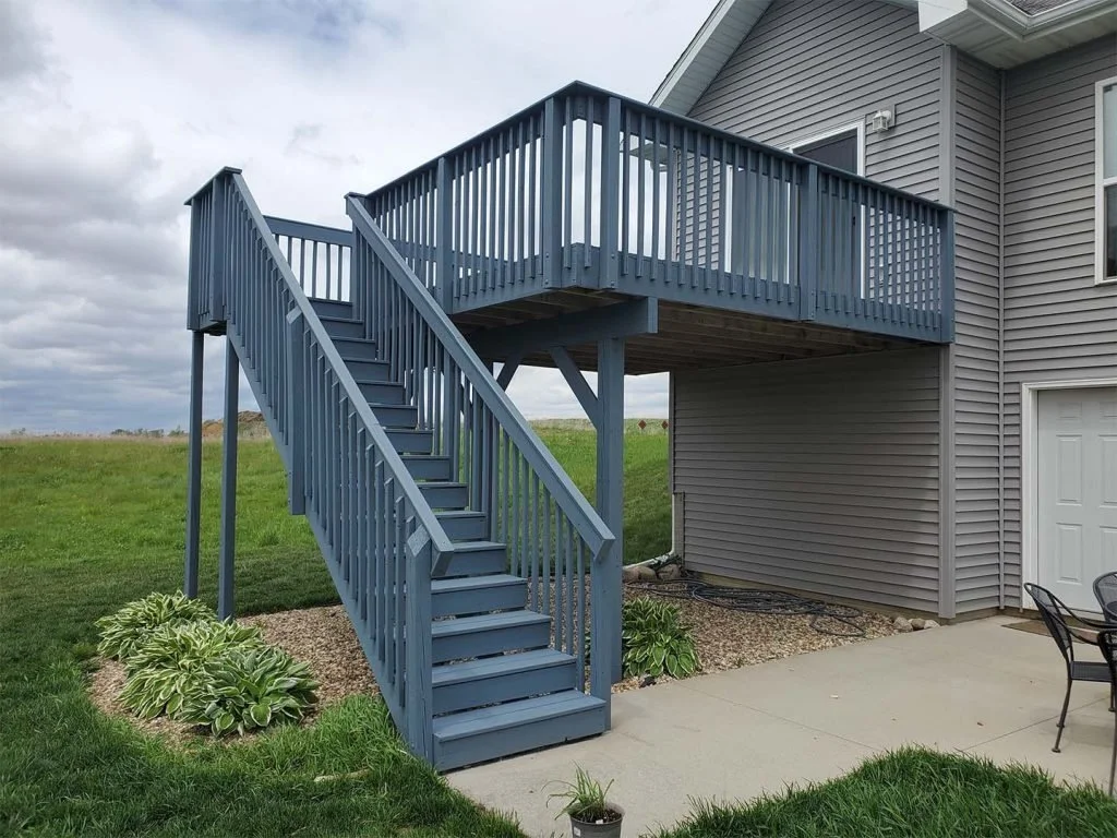 Gray wooden staircase and balcony attached to gray house with outdoor patio furniture and landscaped yard under cloudy sky.