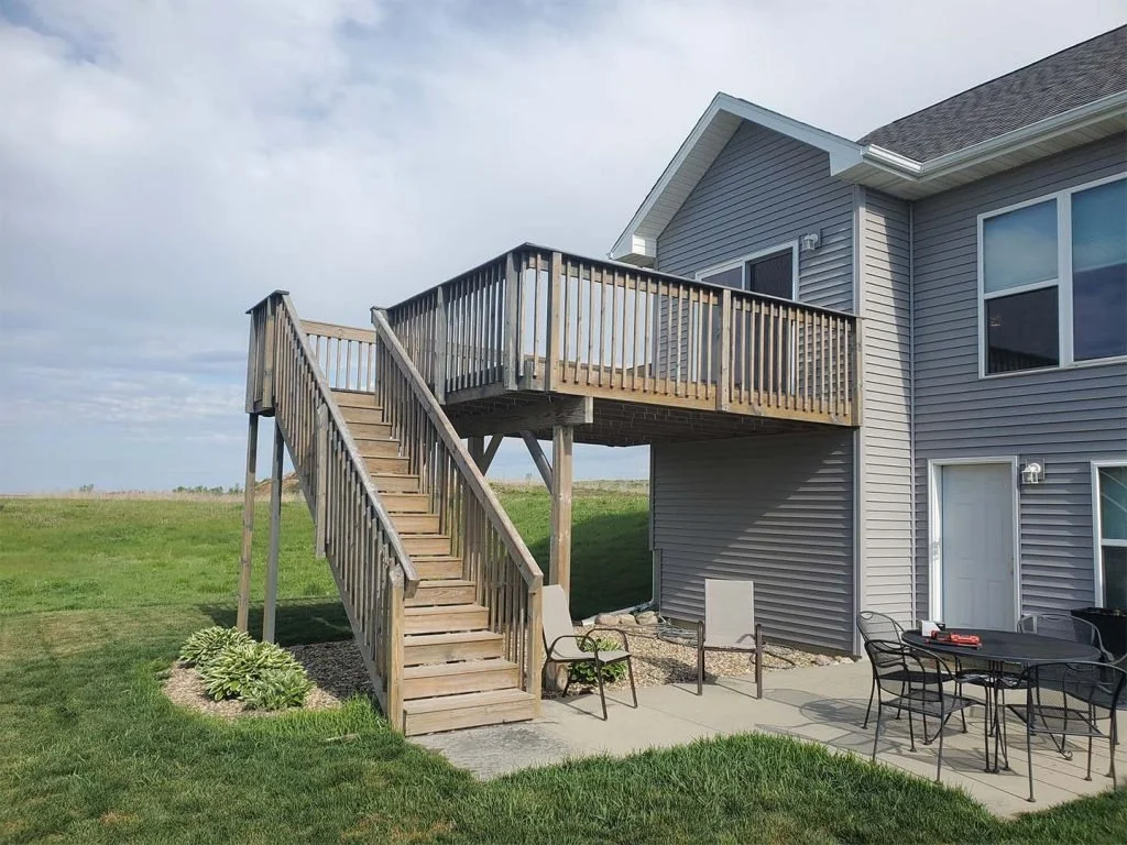 A modern house with gray siding and a black roof, featuring an elevated wooden deck accessible by stairs, with patio furniture below on a concrete slab, and a grassy backyard with an open sky.
