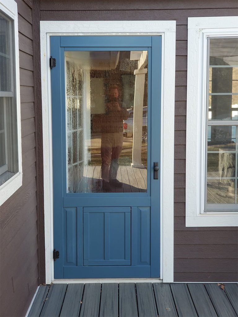 Photo of a blue framed porch door with a glass window, showing a reflection of a person taking the photo, standing on a wooden deck outside a brown house.