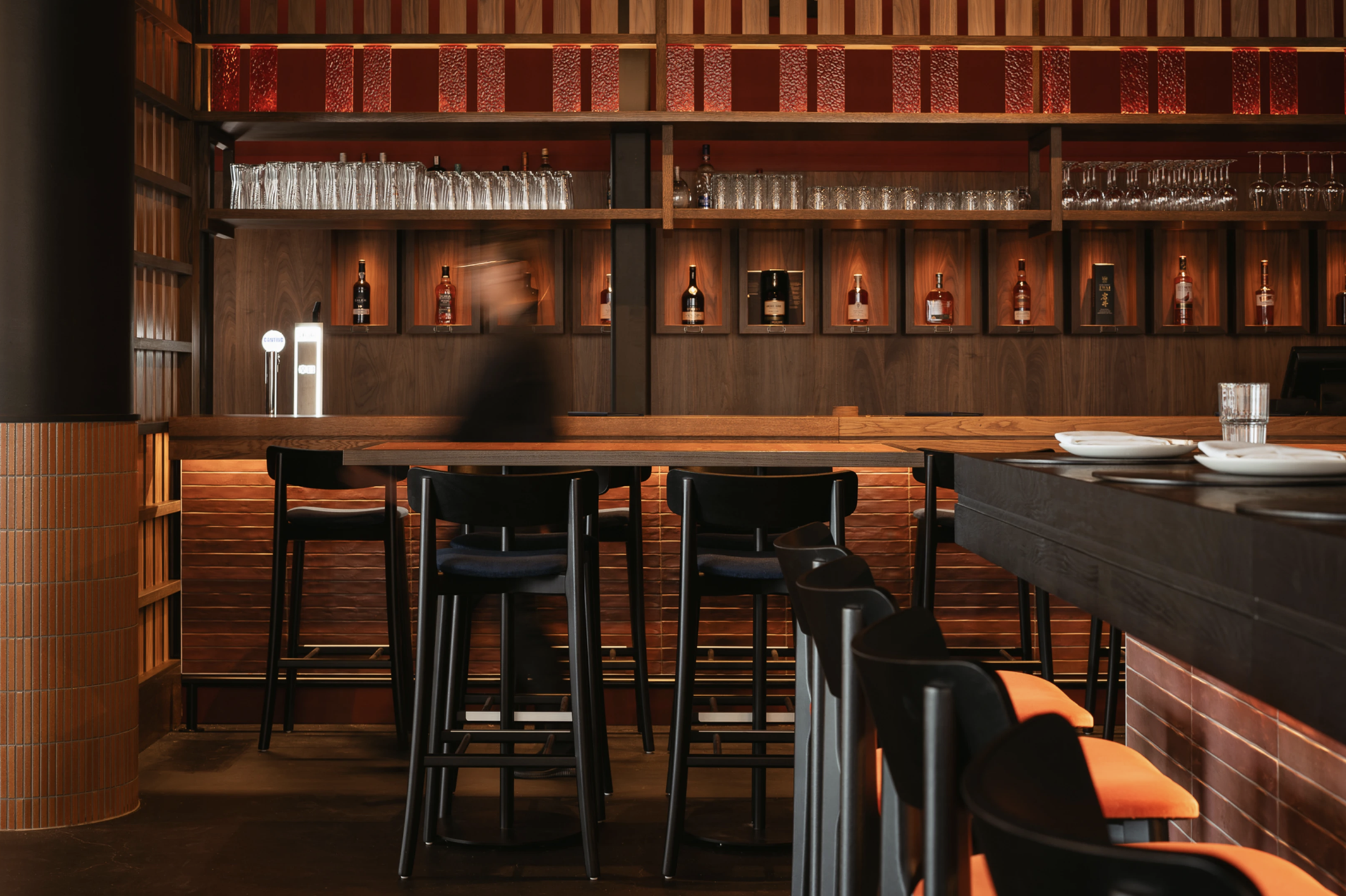 Interior of a bar with wooden shelves stocked with bottles of liquor and glasses, a bar counter with black and orange chairs, and a blurred person in the background.
