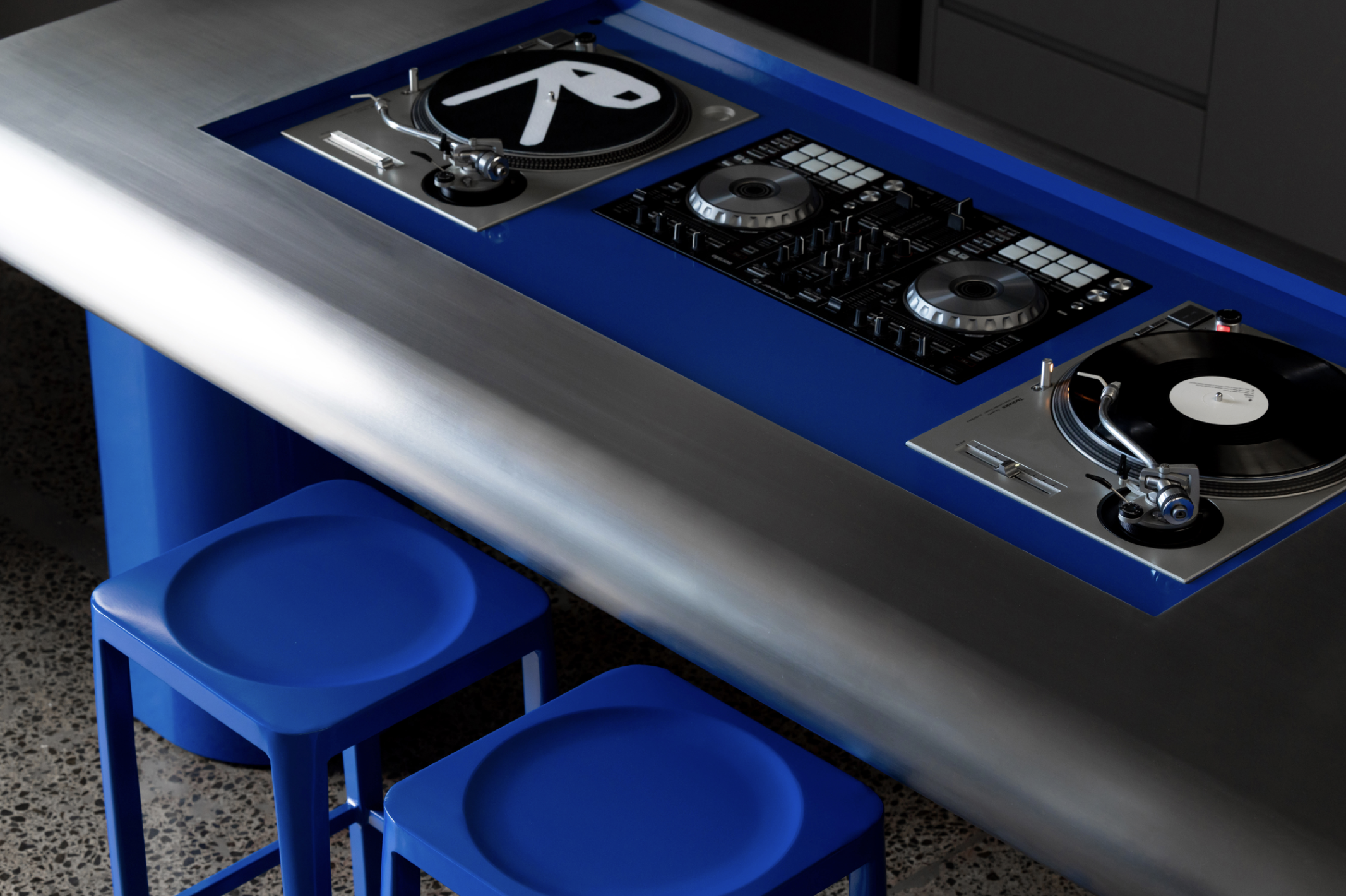DJ booth with turntables and mixer in a modern, minimalistic setting with two blue stools nearby.