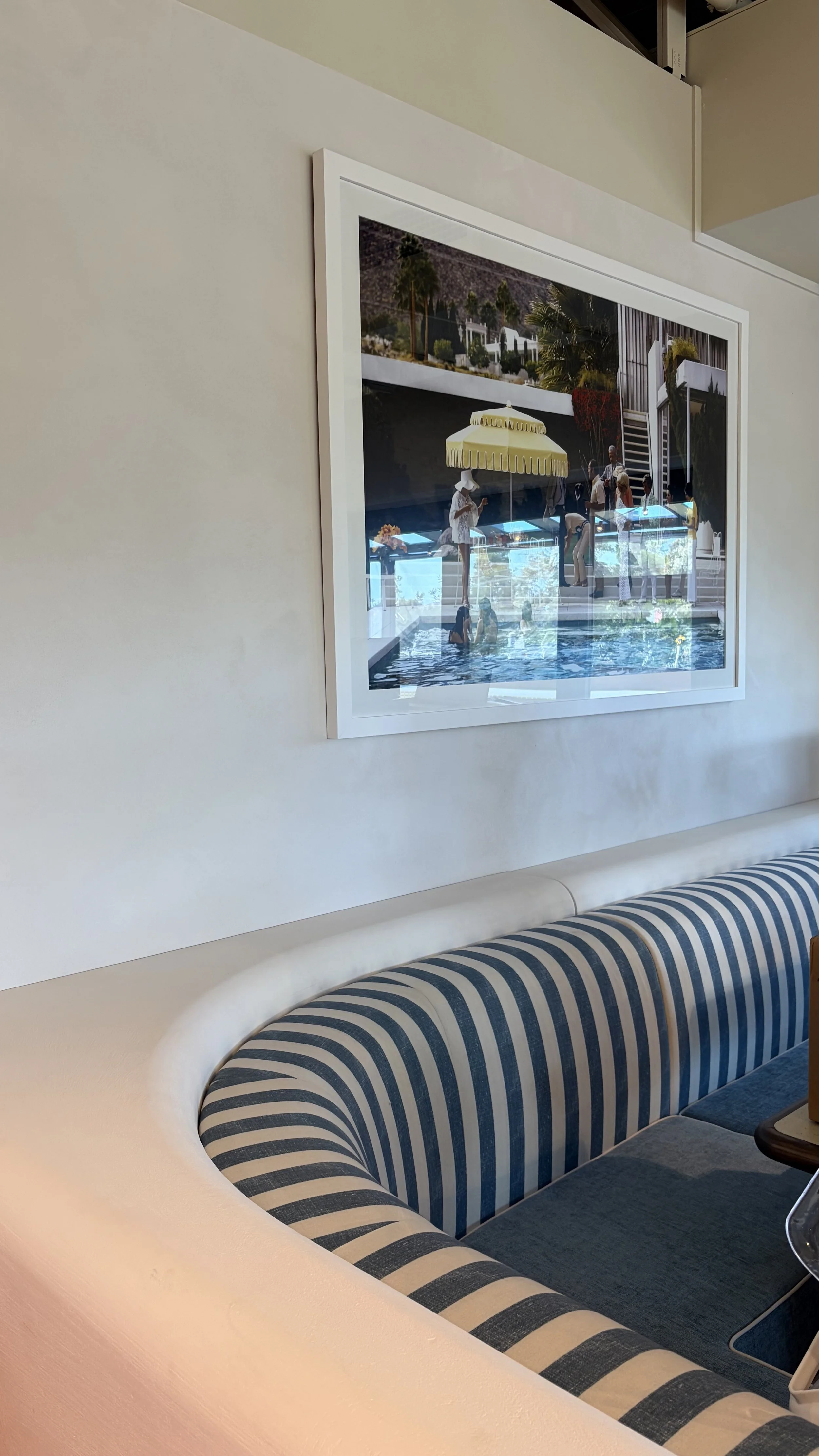 A framed photograph of people relaxing by a pool with a yellow umbrella, mounted on a beige wall above a curved, striped blue and white booth seat in a modern indoor setting.
