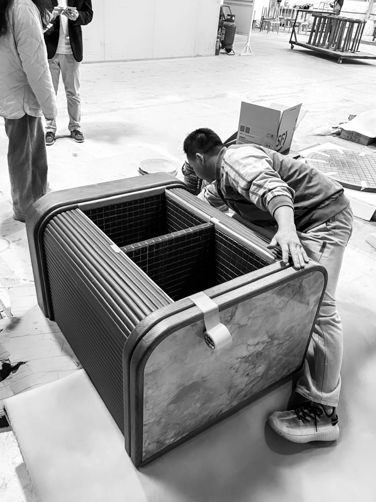 A man in a plaid shirt and sneakers is assembling a modern stylistic rectangular fish tank with a wooden frame. Two other people observe, and the setting appears to be an industrial workspace or showroom with various materials and equipment in the background.