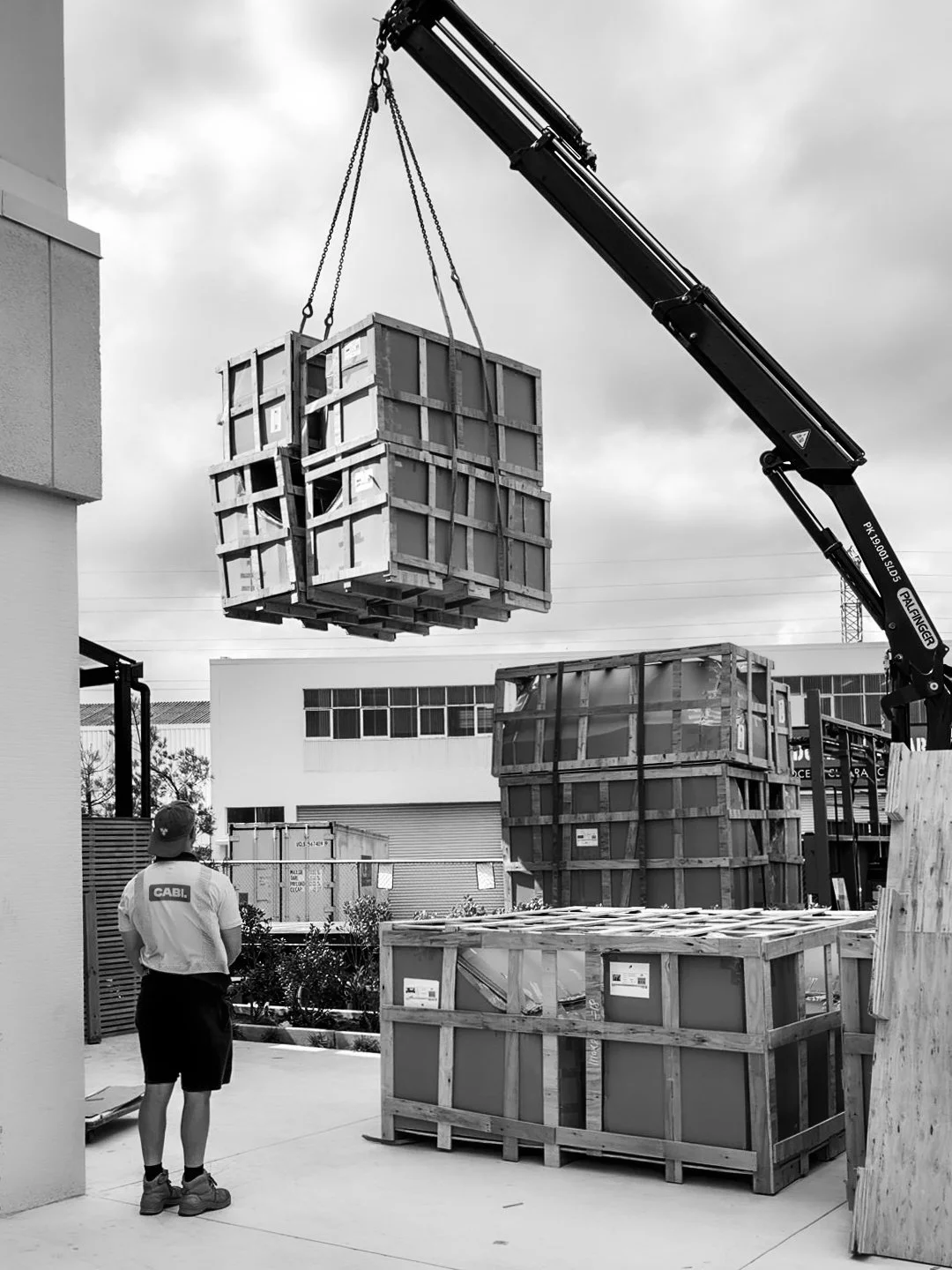 A person wearing a cap and shorts watching as a crane lifts a large wooden crate in an outdoor industrial area.