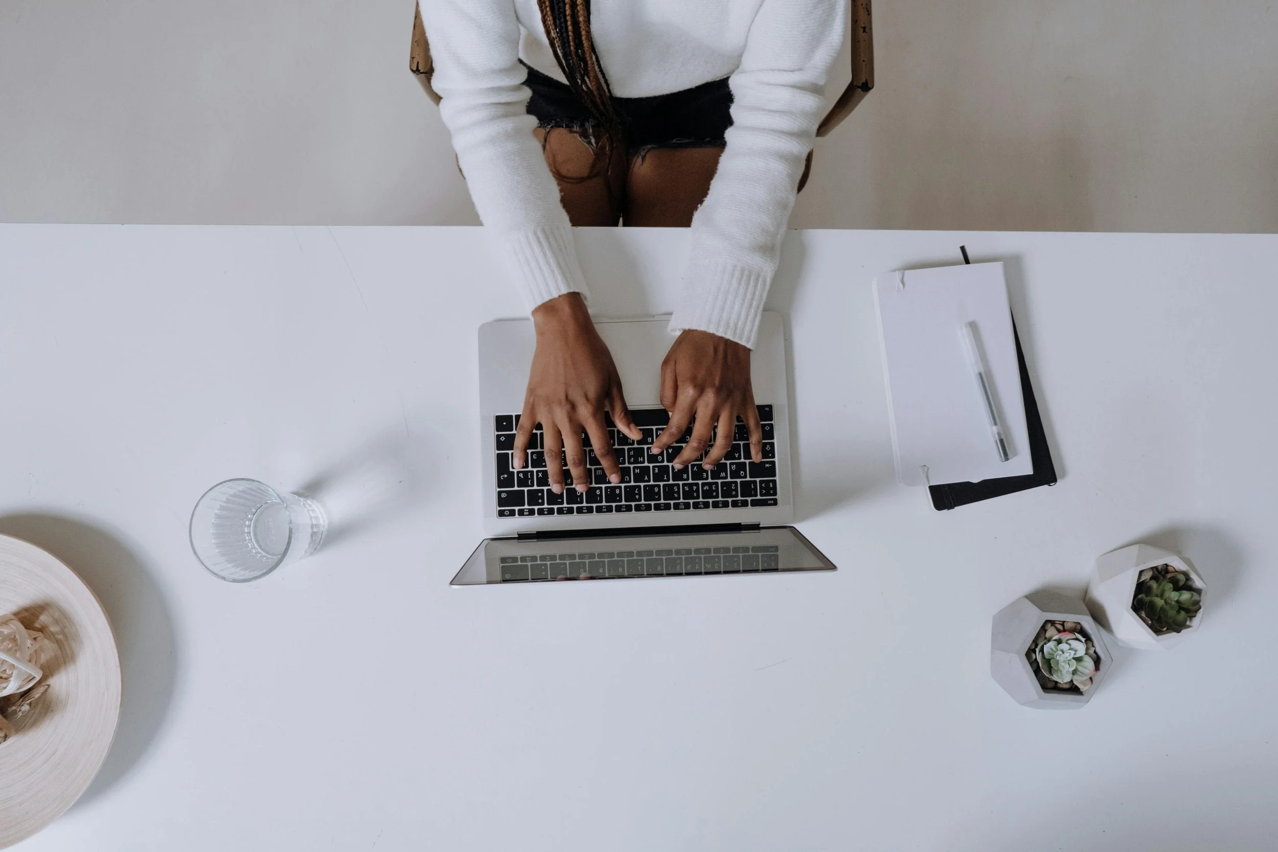 Overhead view of a woman working on a laptop at a clean white desk with a notebook nearby