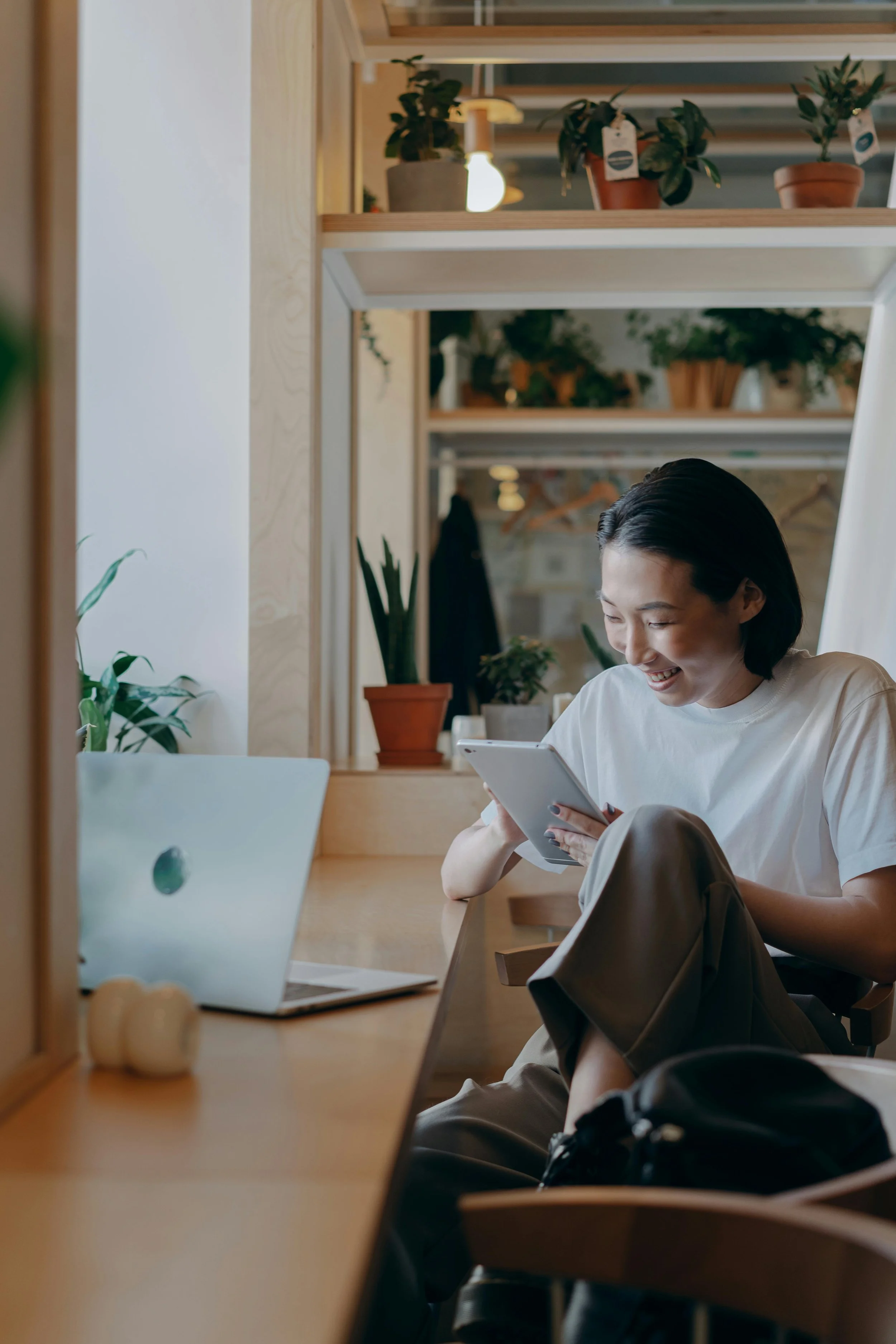 A woman smiling while using a tablet in a bright plant-filled room, representing reaching out to contact a therapist in Meriden, CT at Viridian Counseling
