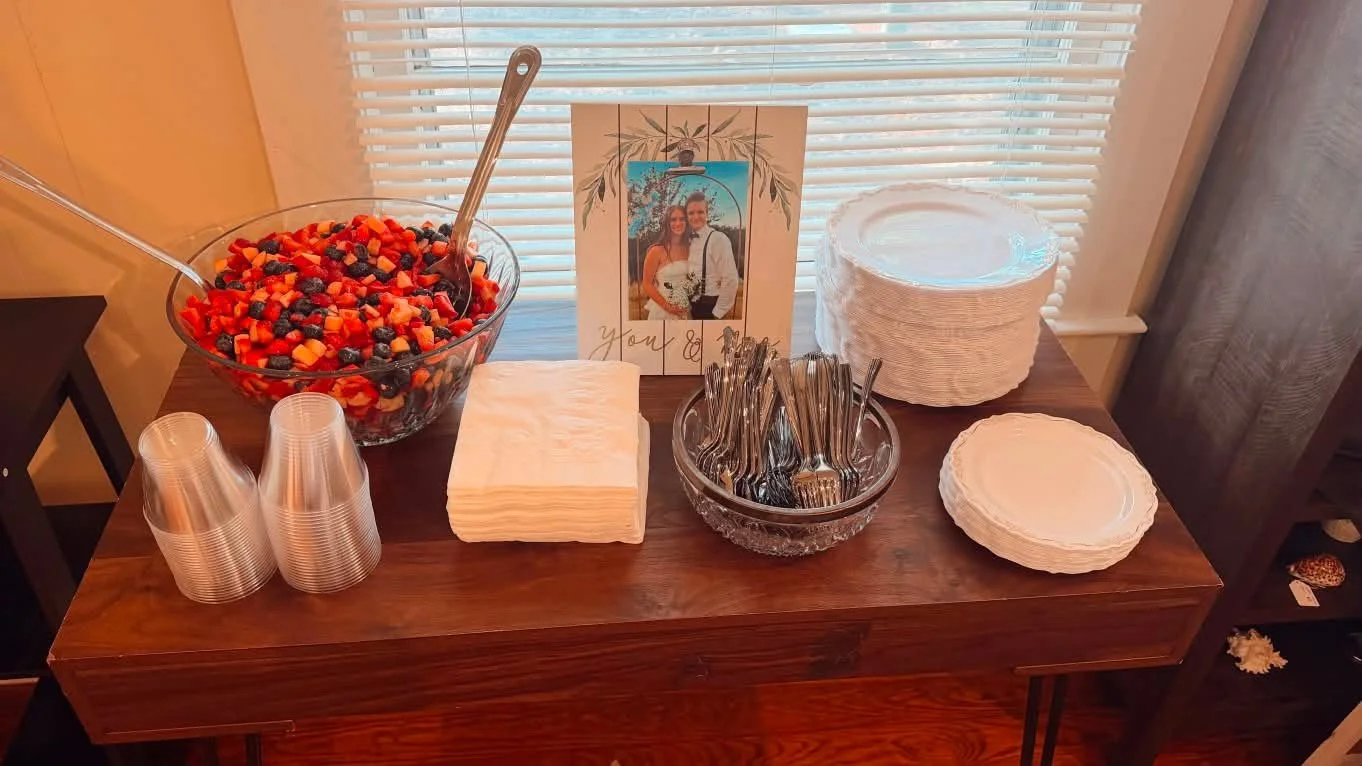 A wooden table set up for a fruit buffet with a large bowl of mixed fruit salad, plastic cups, paper napkins, forks, and plates. A framed photo of a couple with a handwritten message is in the background.