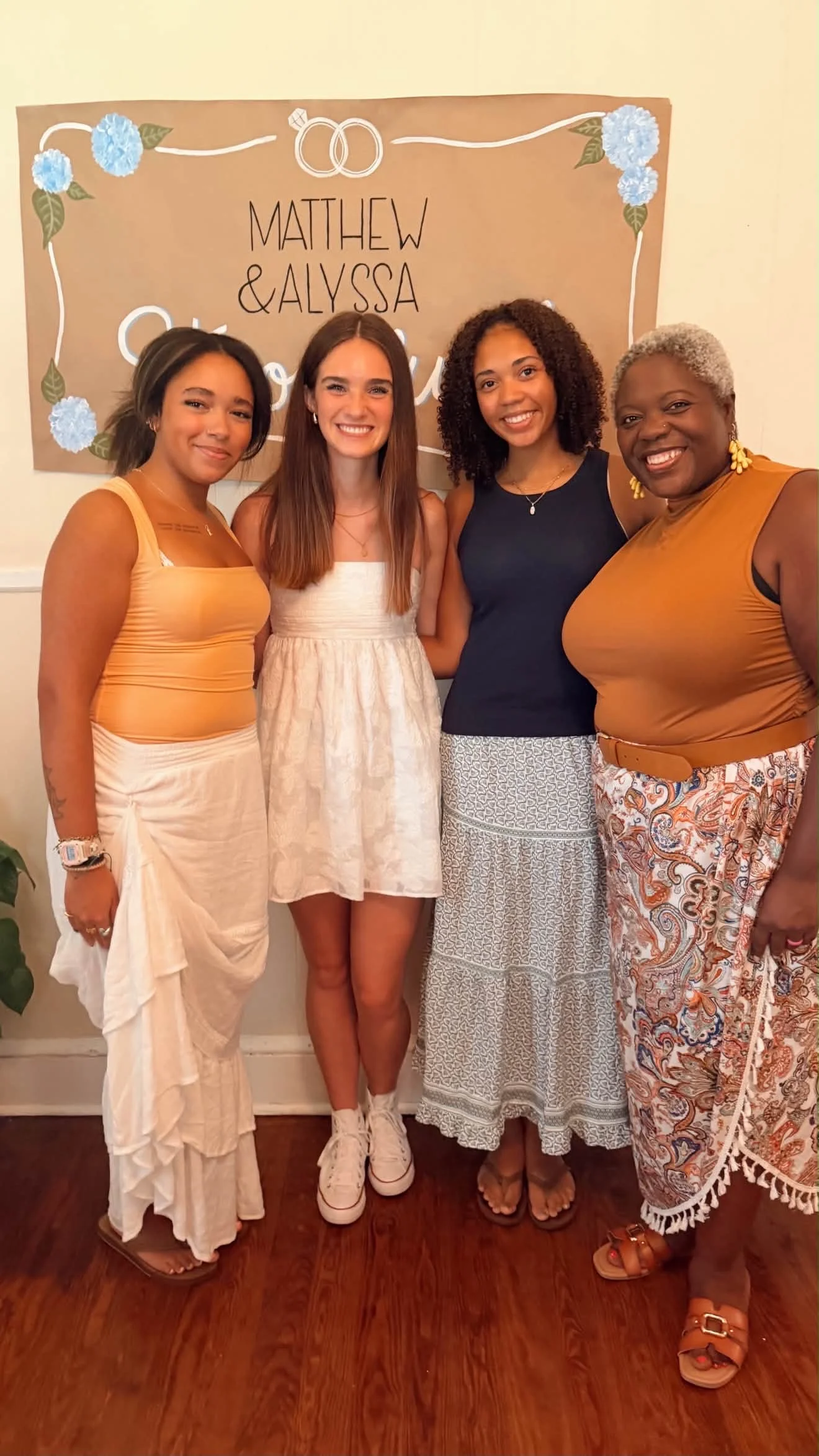 Four women standing together in front of a wedding sign with floral decorations. They are smiling and dressed casually.