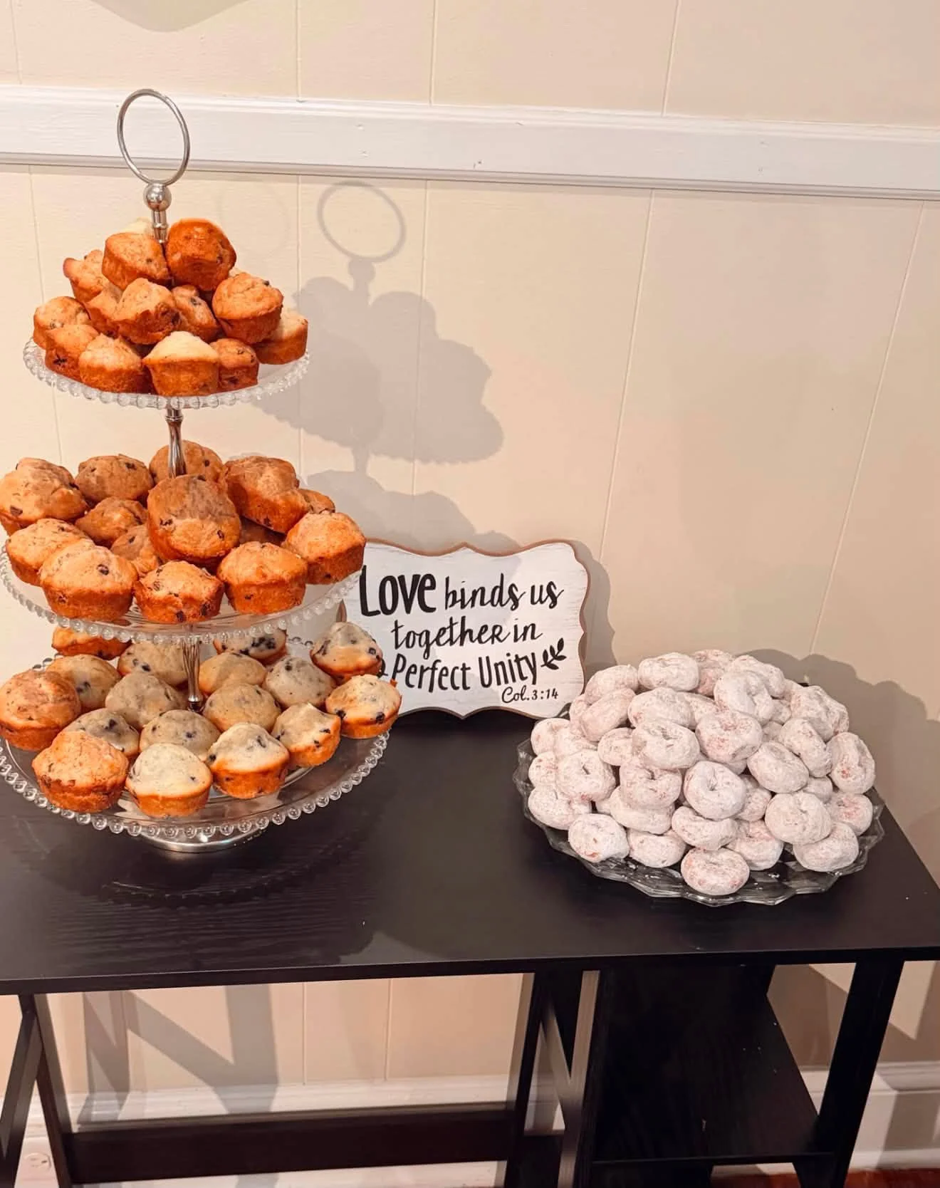 Tiered glass stand holding muffins, some with blueberries, along with powdered donuts and a pile of powdered sugar-covered cookies, with a decorative sign that reads 'Love binds us together in perfect unity' against a beige tiled wall.