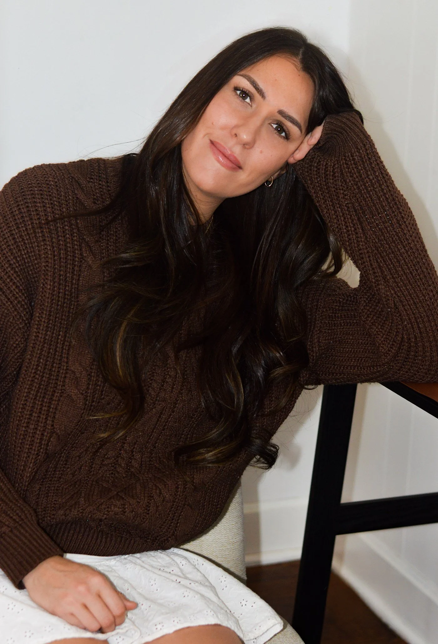 A woman with long dark hair, wearing a brown sweater, sitting on a beige chair, resting her head on her hand, next to a black table, indoors against a white wall.