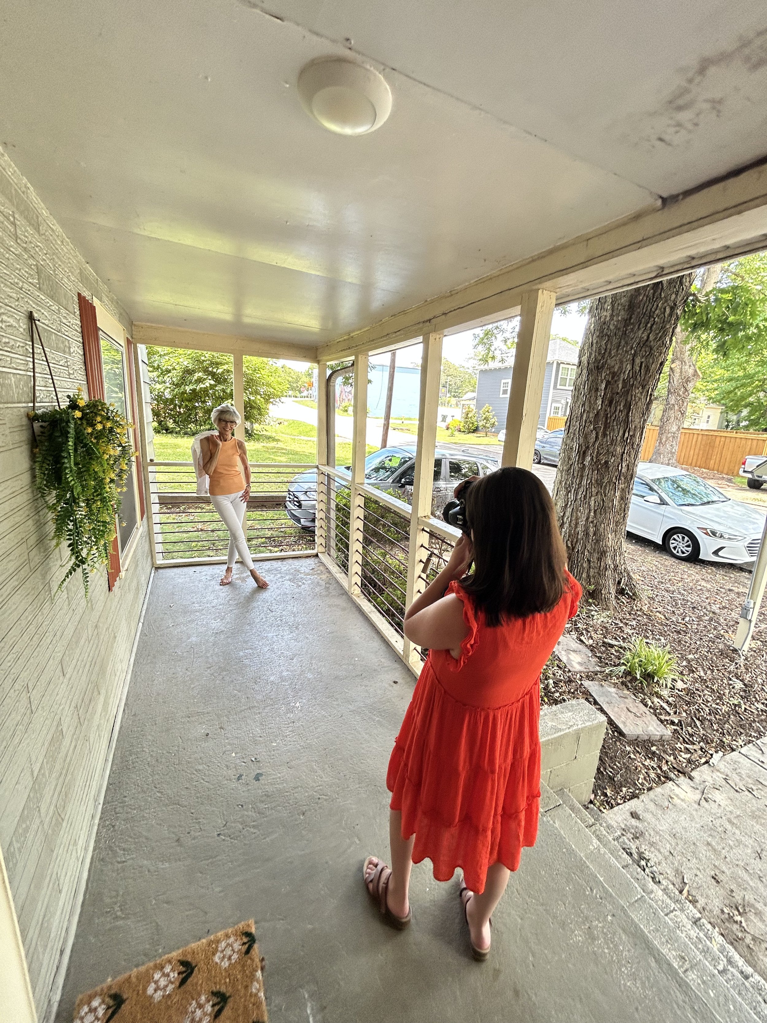 A woman with gray hair, glasses, wearing an orange sleeveless top and white pants, standing on a porch, smiling and talking to another woman with brown hair, wearing a red dress, who is facing away from the camera.