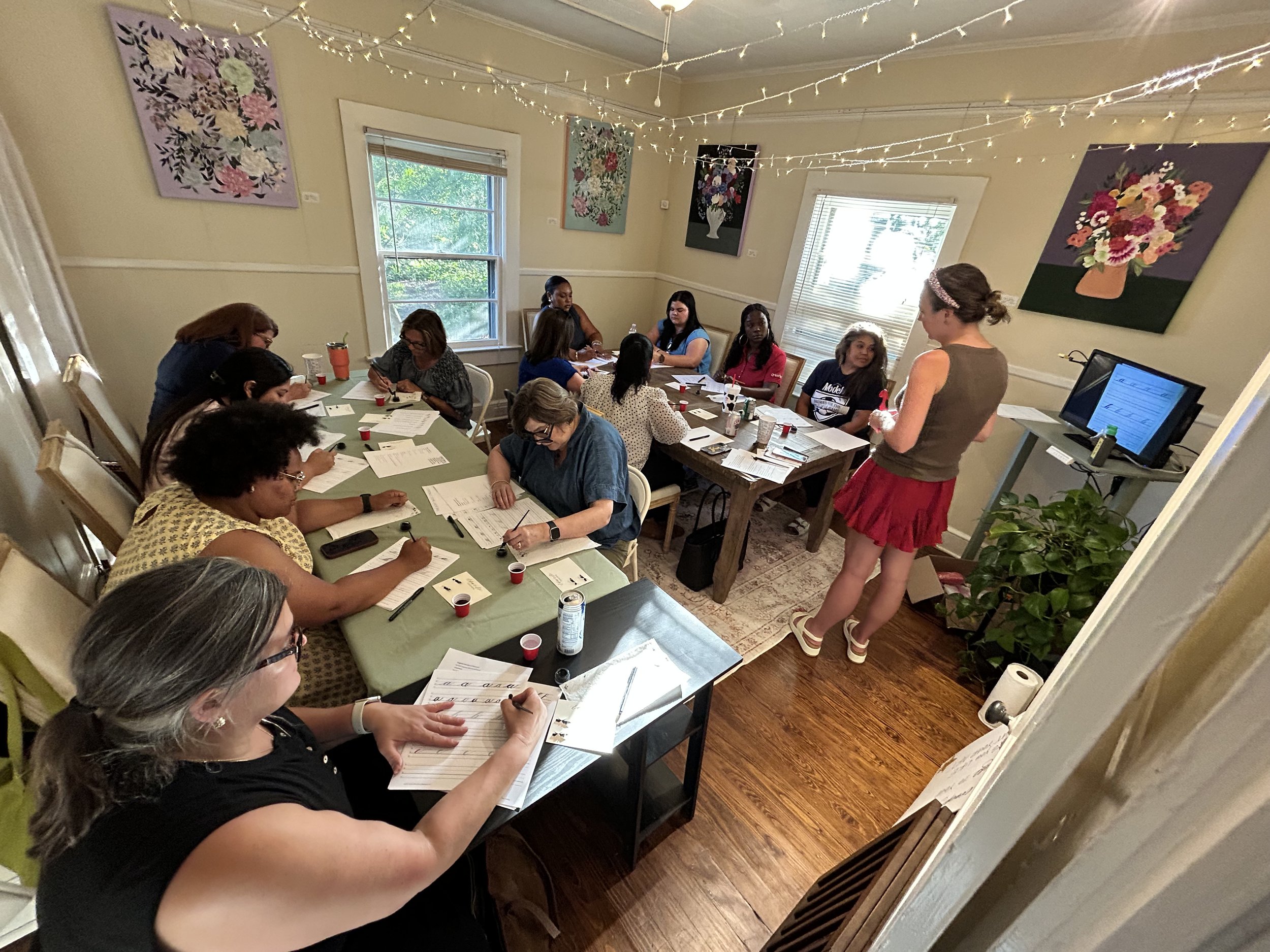 A group of women participating in a calligraphy or handwriting class in a decorated cozy room with paintings of flowers on the walls and string lights hanging from the ceiling.