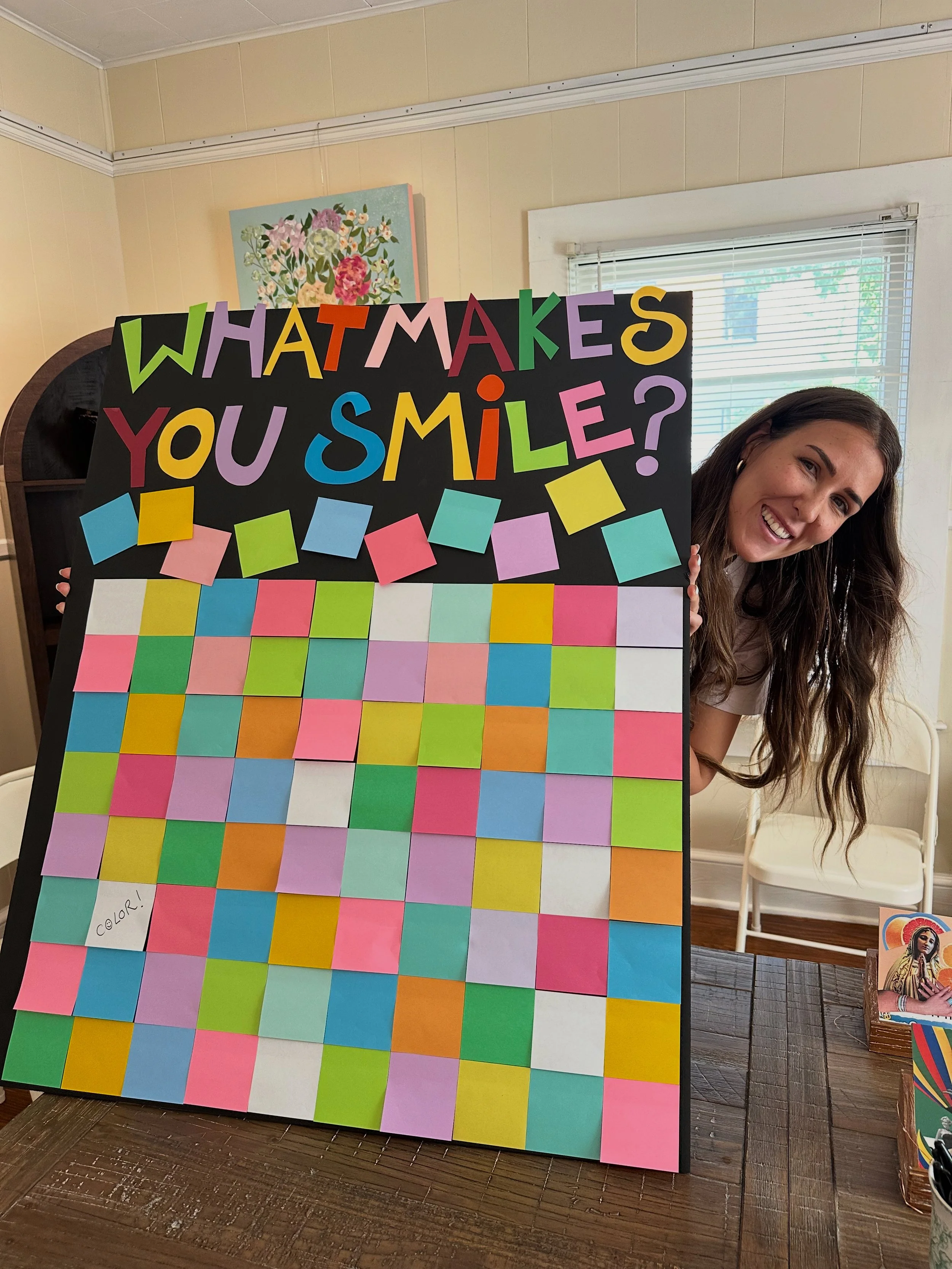 A woman smiling and holding a colorful poster board with the question 'What makes you smile?' written on top. The board is decorated with multicolored sticky notes arranged in a grid pattern.