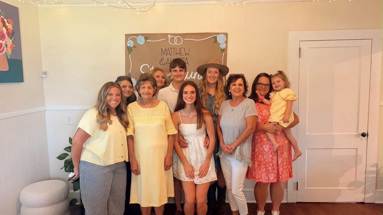 Group of ten people, including children and adults, gathered indoors for a celebration, standing in front of a brown backdrop with white and blue decorations and handwritten text. Everyone is smiling.