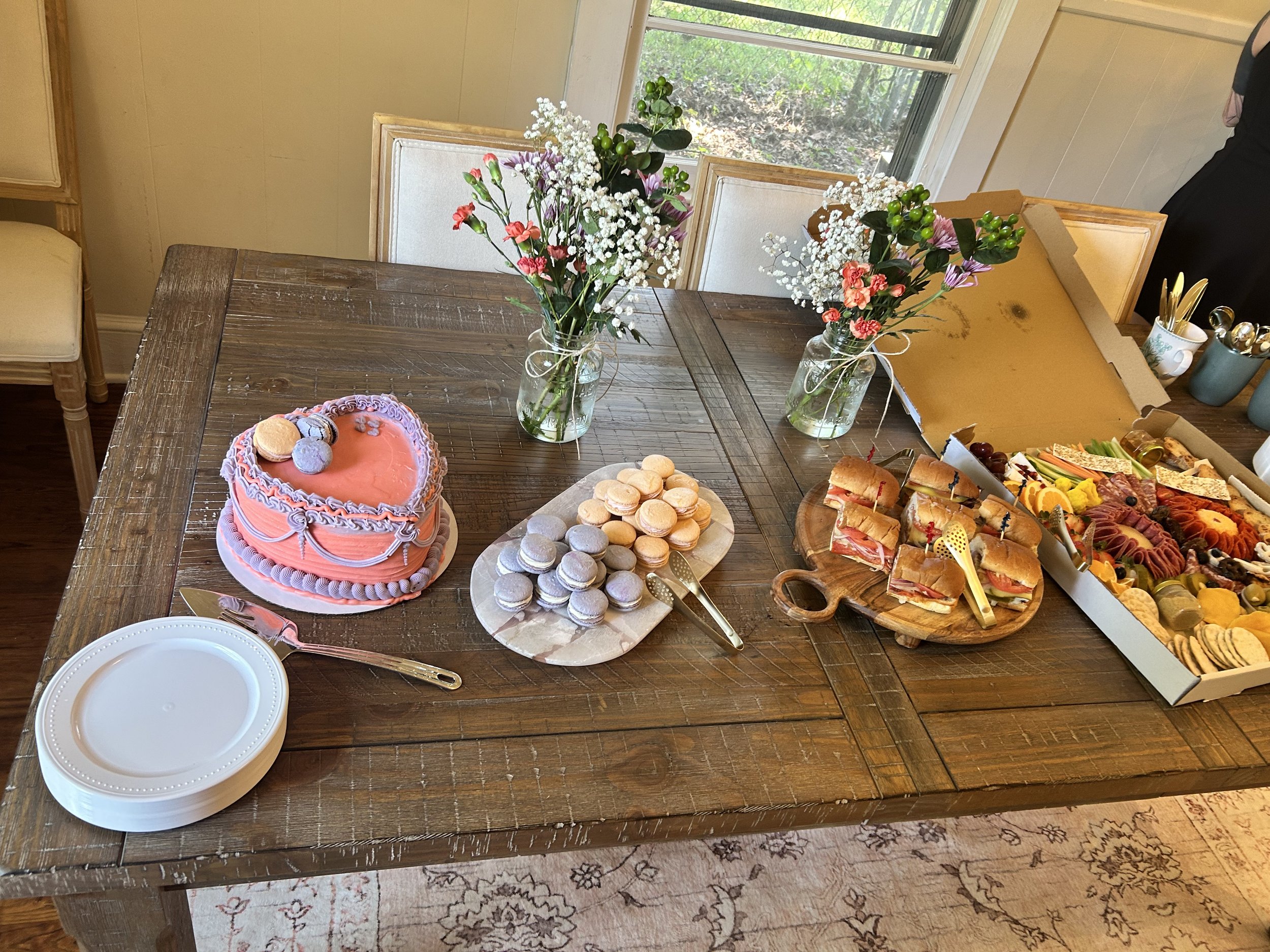 A wooden table set with a heart-shaped pink cake, macarons, and a platter of finger foods, with flowers in vases and a window in the background.