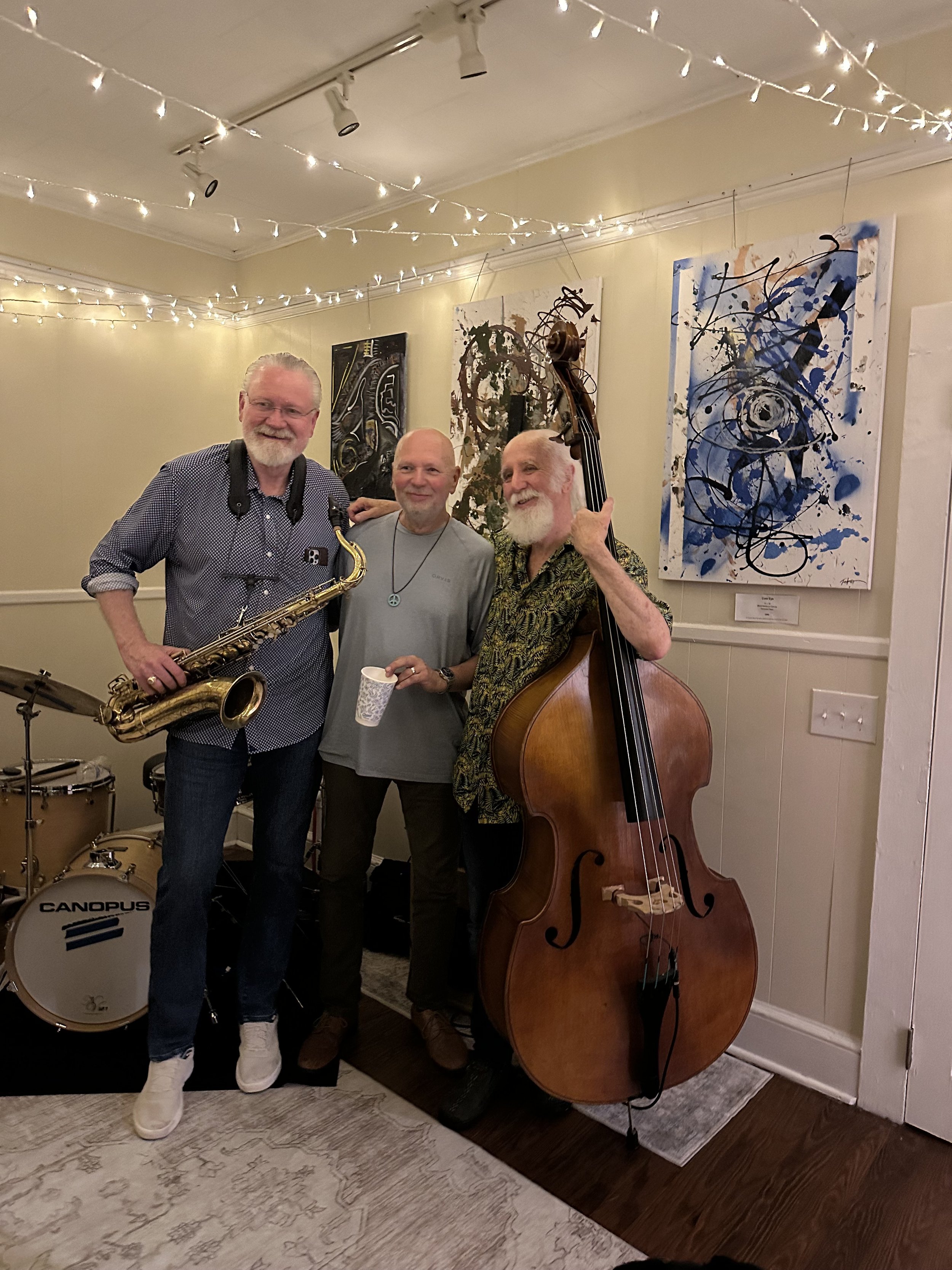 Three men standing together at a music event in front of abstract paintings on a wall with string lights overhead. One man is holding a saxophone, another an upright bass, and a drum set is visible in the background.