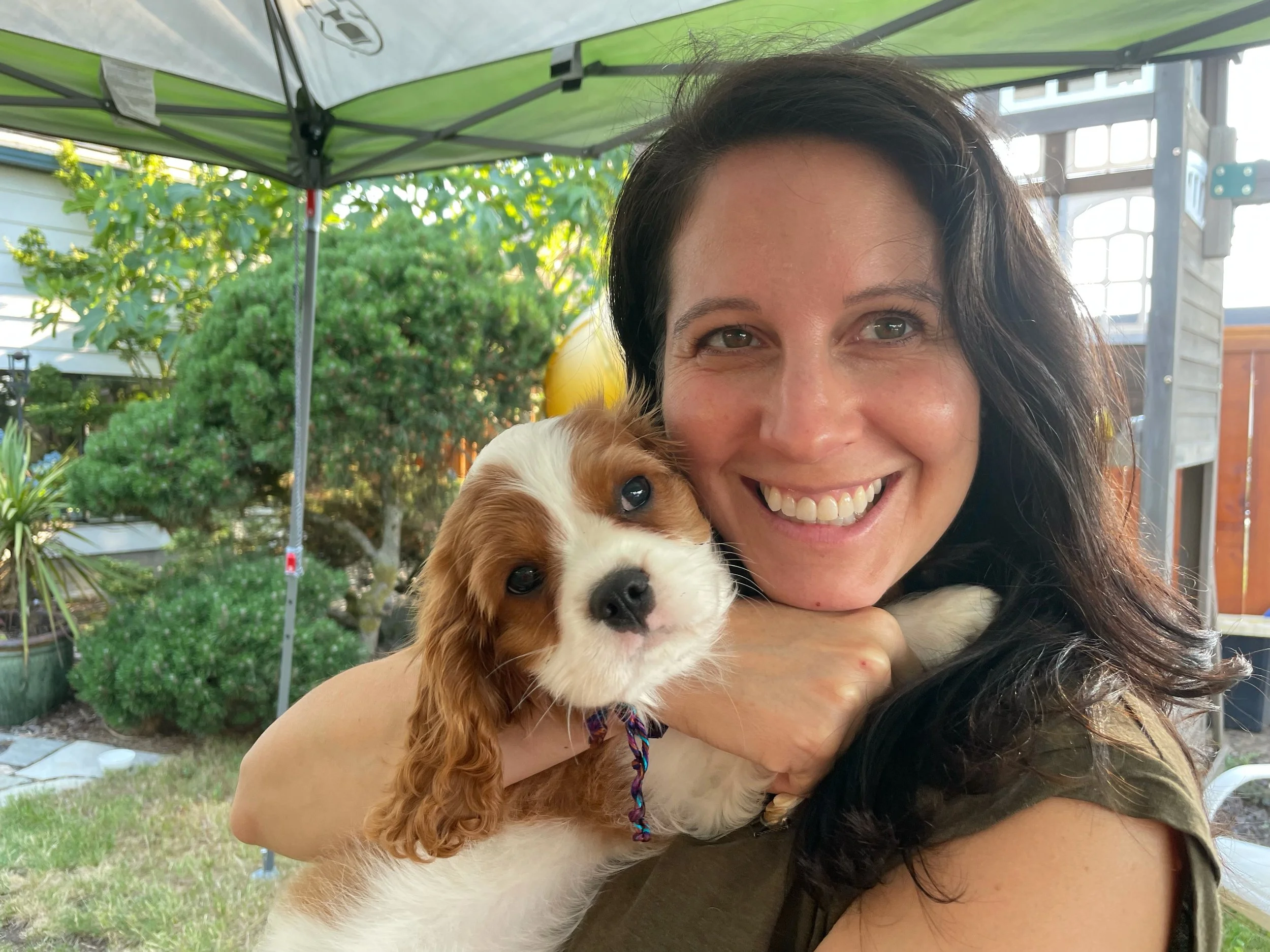 A woman smiling and cuddling a cute puppy outdoors in a backyard with a green tent canopy overhead, trees, and garden decorations in the background.