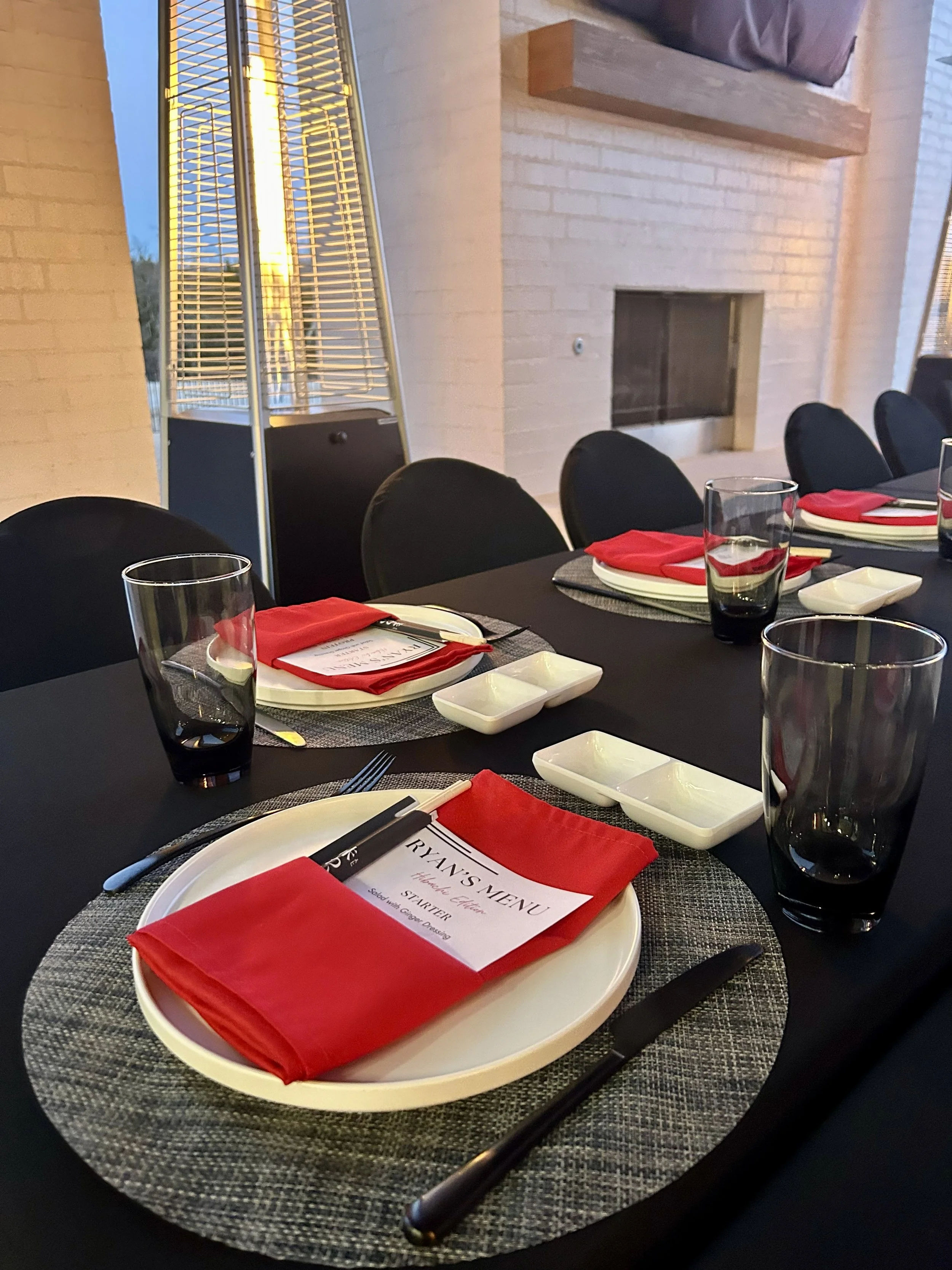 A dining table set for a meal with black chairs, white plates, red napkins, black cutlery, and clear glasses, arranged on black tablecloths with round gray placemats on an outdoor patio for a Hibachi birthday Party.