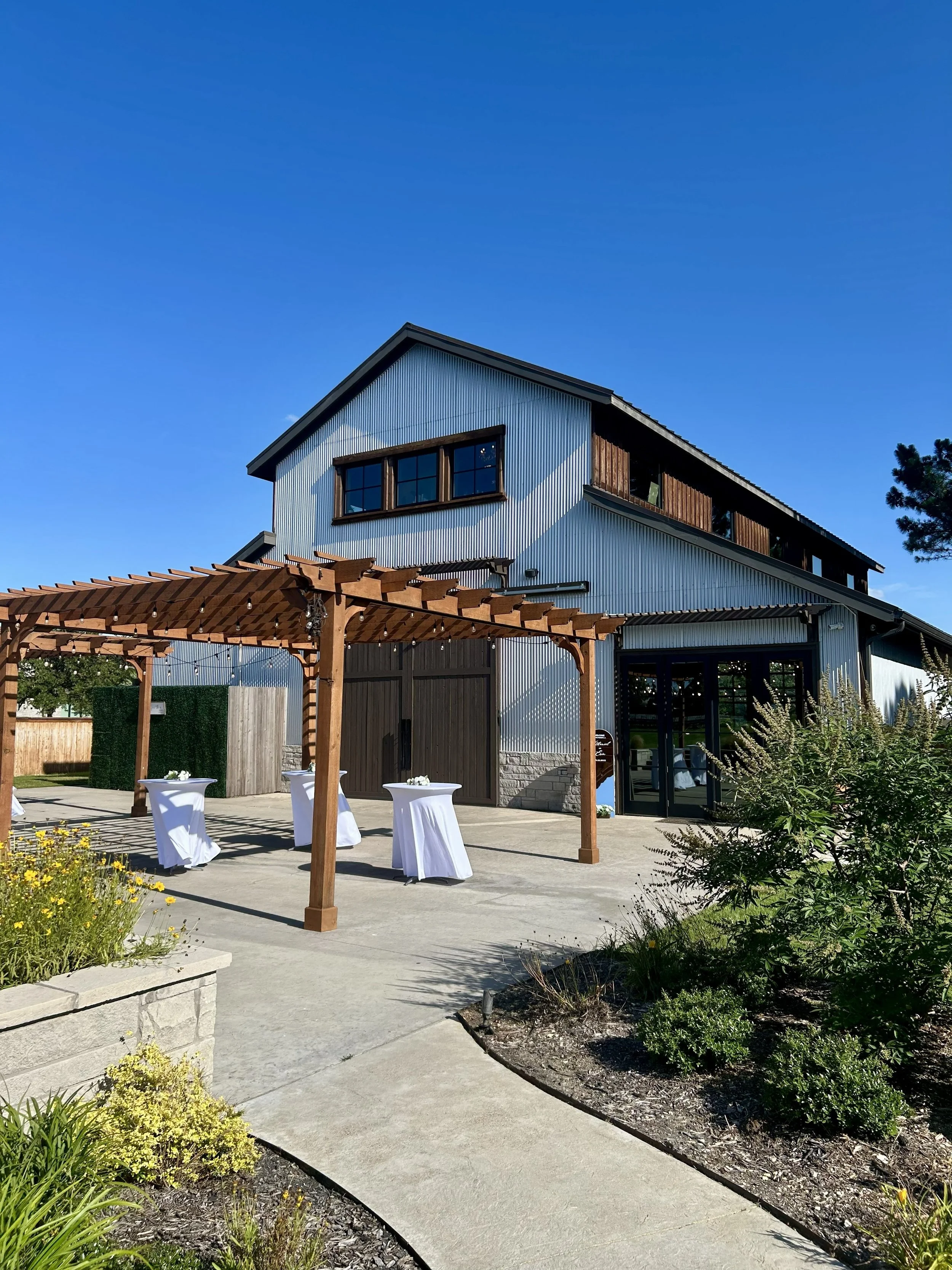 An industrial barn wedding venue with a metal exterior and wood accents on the top floor, with a wooden pergola and high-top tables with white tablecloths outside, surrounded by greenery and a clear blue sky.