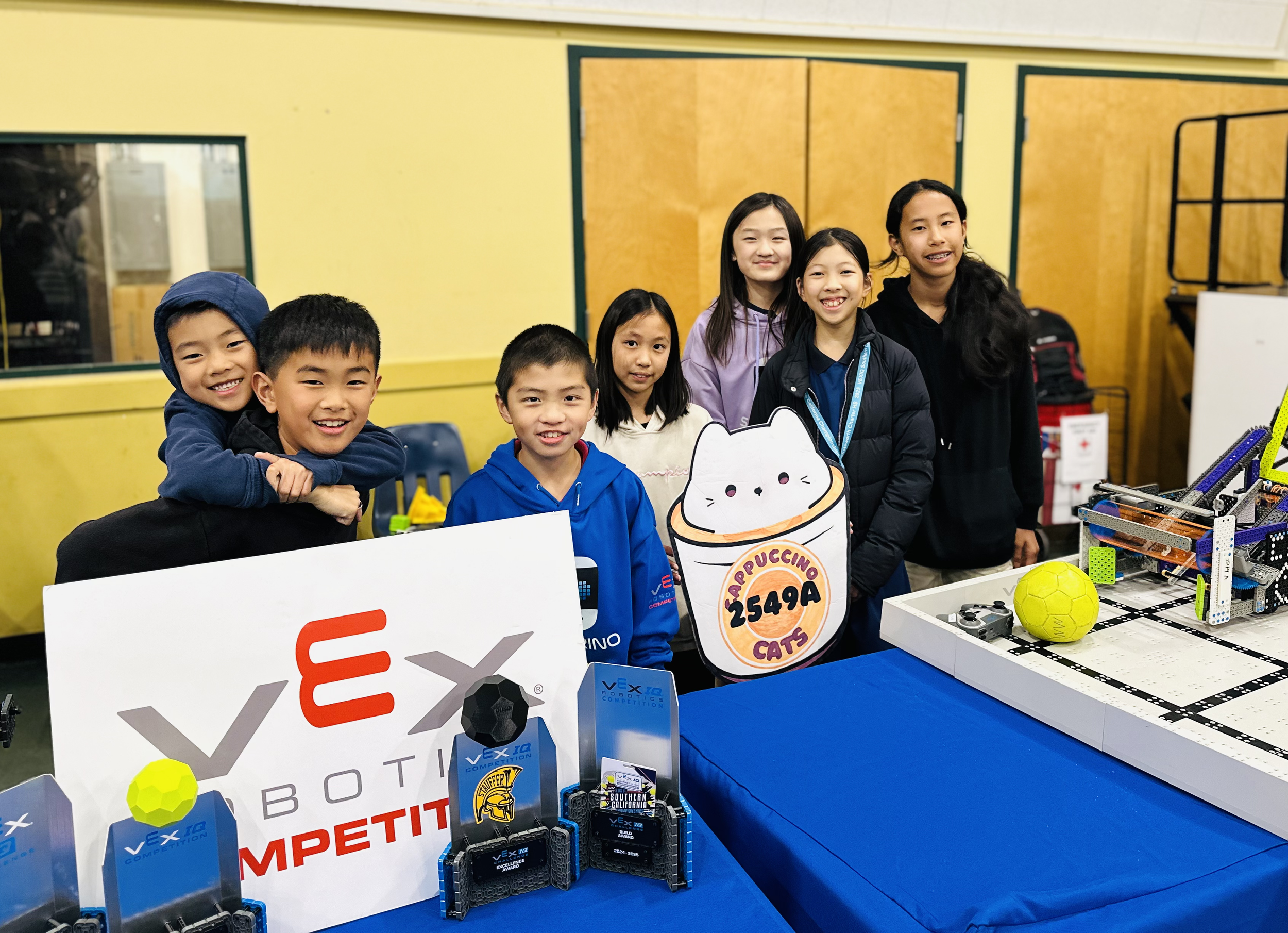 Group of children at a robotics competition booth, smiling, with trophies and robot parts on the table, yellow wall in the background.
