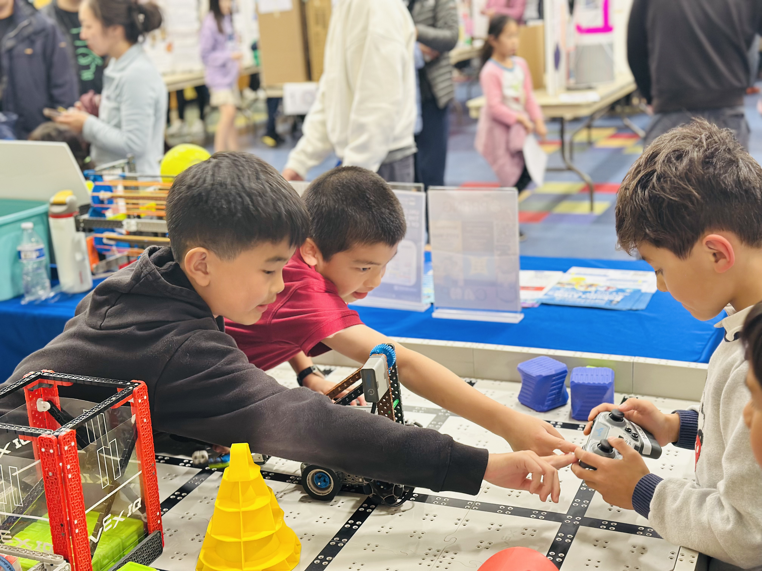 Children and adults at a STEM exhibition, working with robotics and technology projects on a table in a large indoor space.