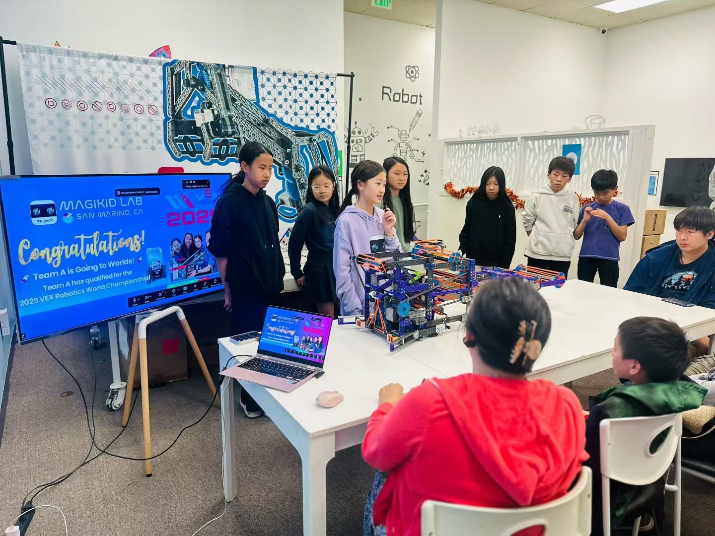 Group of students and adults in a classroom around a robotics project, with a large display screen showing a message about a robotics competition.