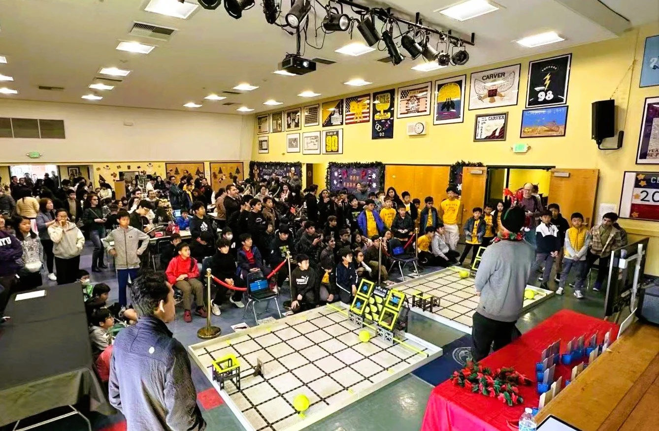 Large indoor robotics competition with many students and spectators, a table with robot matches, and decorated walls.