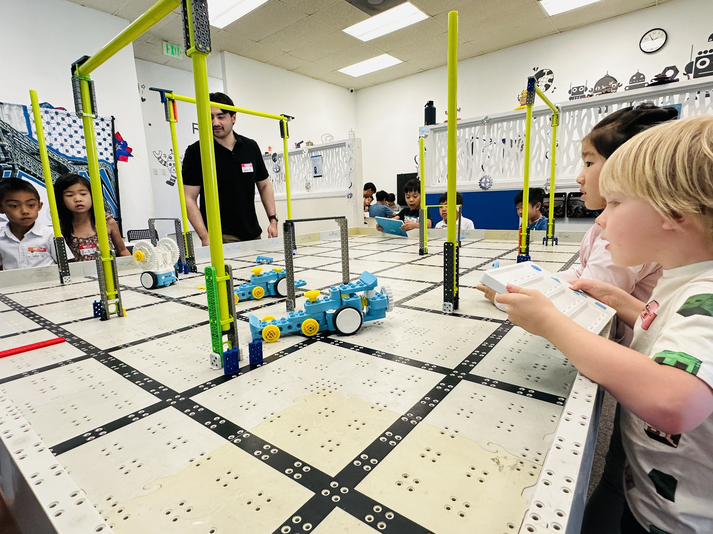 Children and adults watch and operate a LEGO robotics competition on a white table with black grid markings. The scene is inside a classroom with decorative wall art, a clock, and fluorescent ceiling lights.