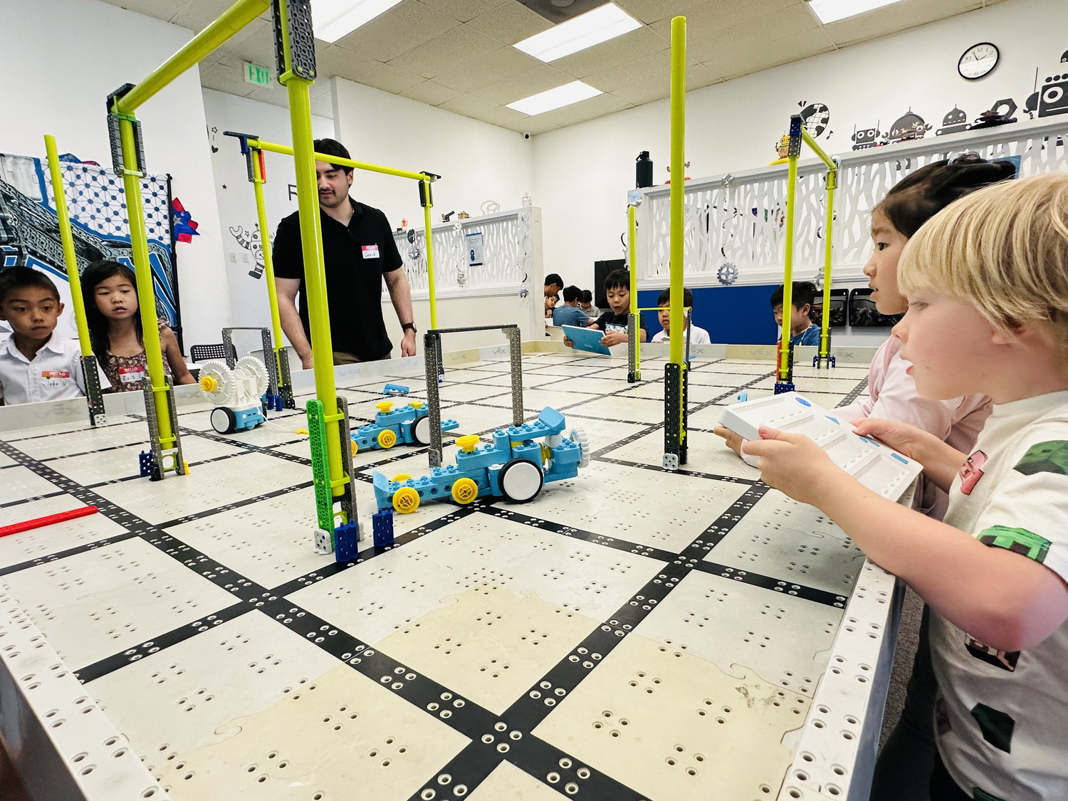 Children and adults operating a LEGO robotics competition on a white table with black grid markings