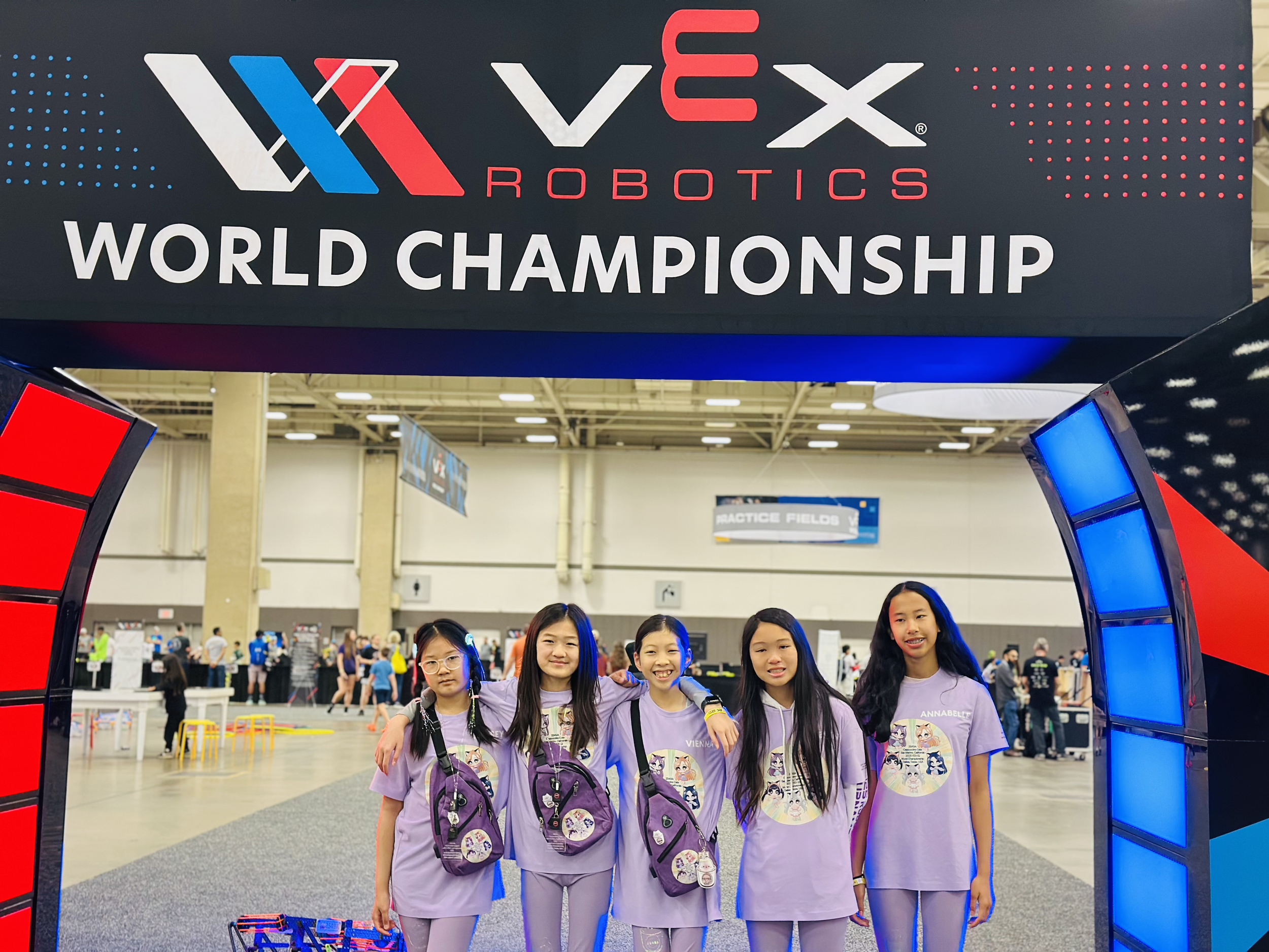 A group of five young girls wearing purple T-shirts with cartoon character graphics, standing inside a large arena at the VEX Robotics World Championship. They are smiling and posing for the photo, with some carrying purple backpacks. In the background, there are other participants, event booths, and banners.