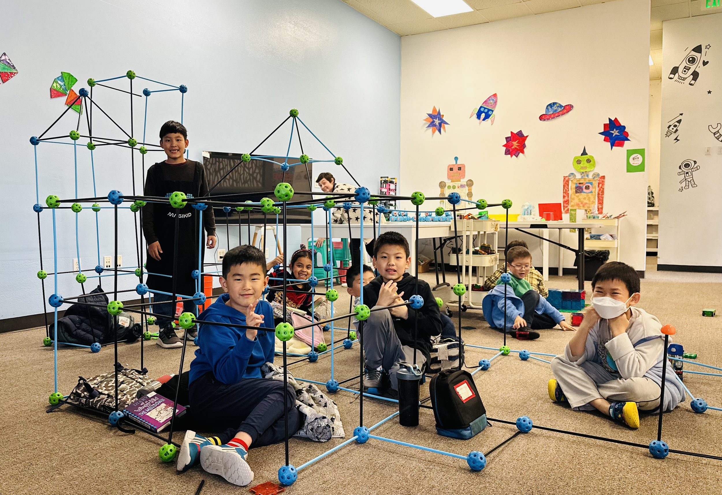 Children sitting on the floor in a classroom, surrounded by educational toys and decorations, with several children smiling and a boy standing behind a large plastic geometric structure.