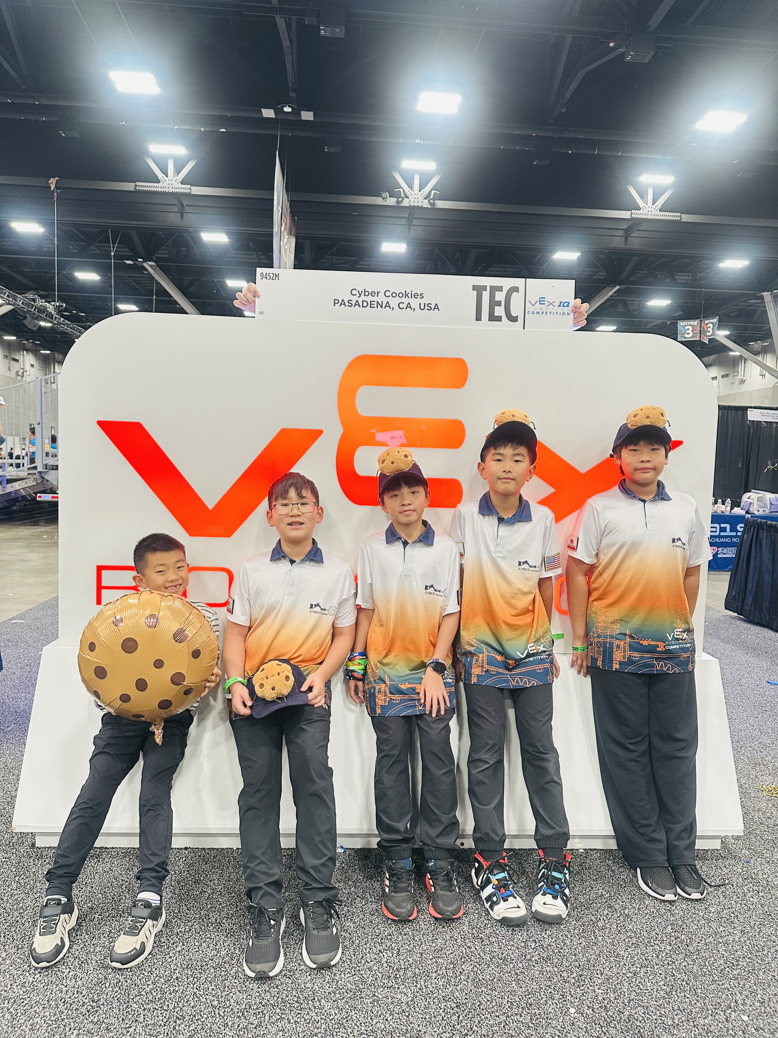 A group of six young boys standing in front of a white display board at an indoor robotics event. They are dressed in matching team shirts with a gradient orange to navy design, and some are wearing black caps with plush cookies attached. One boy on the left is holding a large cookie-shaped balloon. Behind them, a sign reads 'Cyber Cookies, Pasadena, CA, USA'. The setting appears to be a convention or competition with industrial lighting and booths in the background.