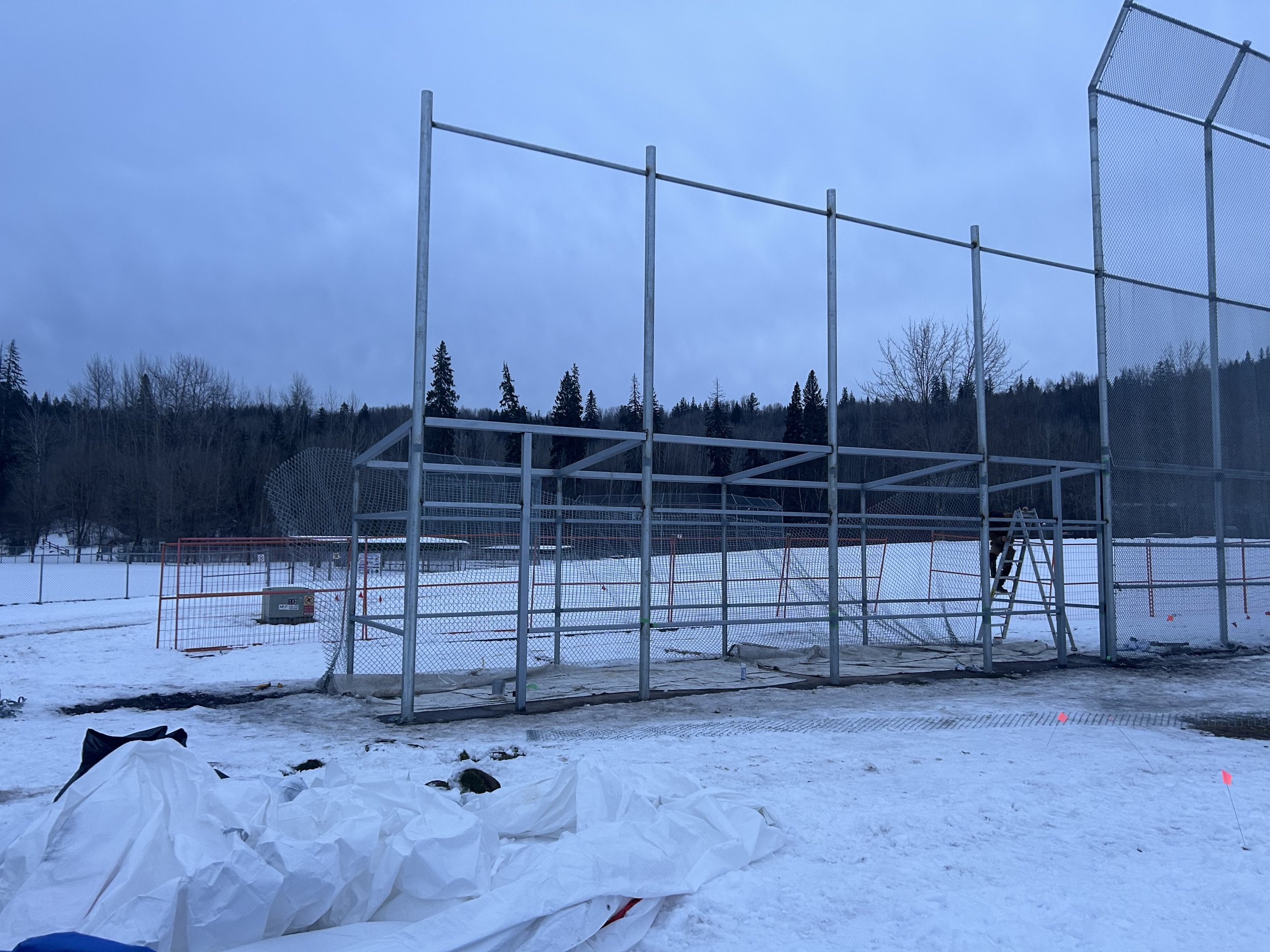 Construction site in snowy outdoor area with metal framework and chain-link fencing, ladder, and construction materials.